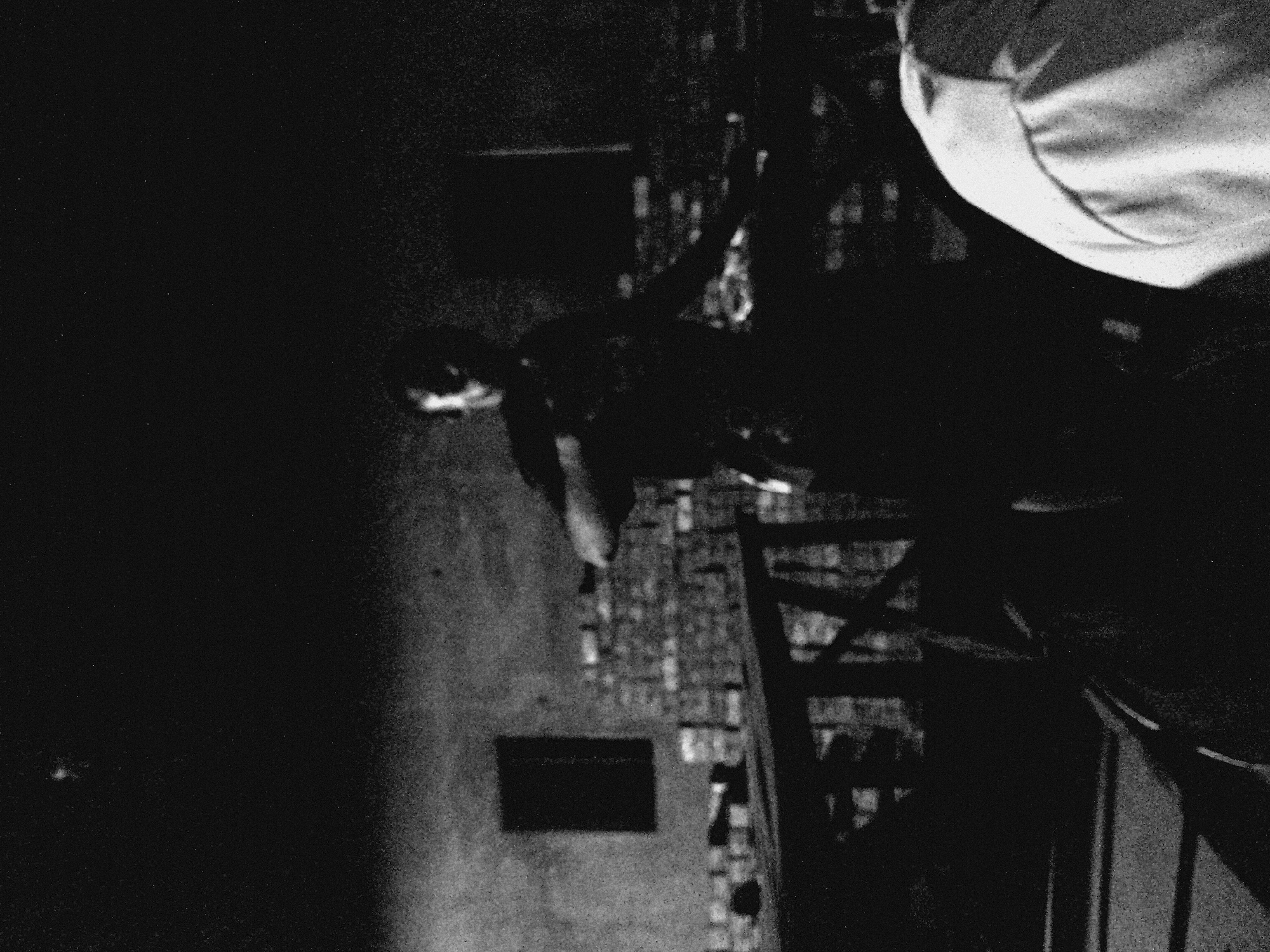 The image depicts an individual standing in a seemingly urban environment. The individual is wearing what appears to be a light-colored shirt or garment. The background includes what could be a brick pathway and a dark, indistinct wall. The scene is dimly lit, making details difficult to discern. The location is Unknown, Unknown.