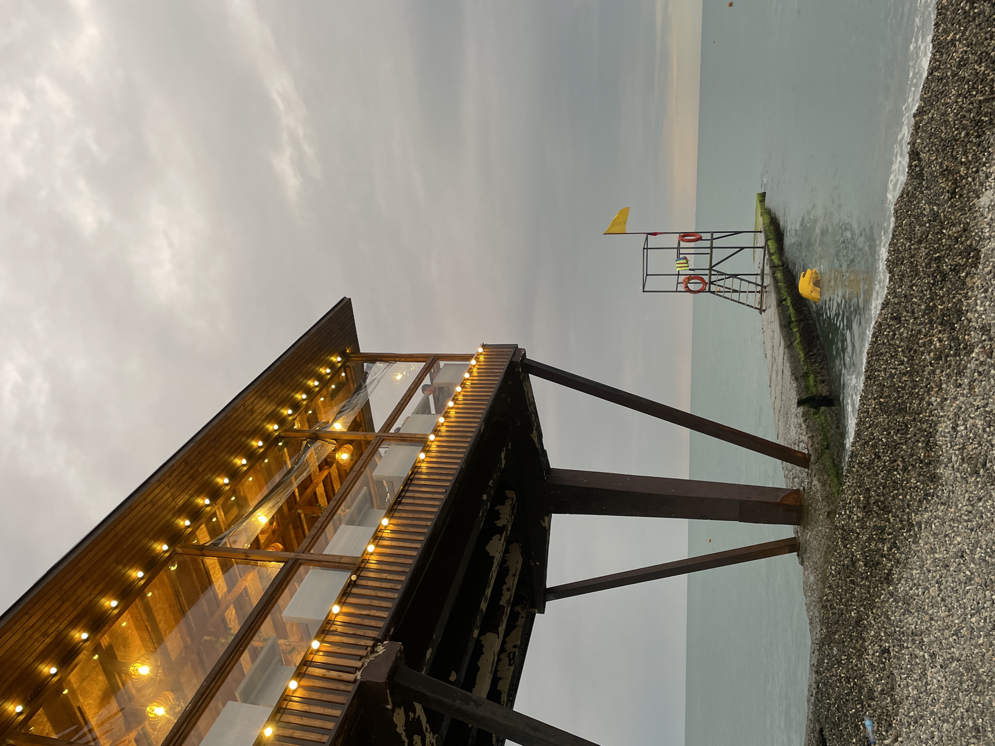 The image shows a pier extending into a calm, gray sea under an overcast sky. On the left, a wooden structure with a transparent roof and strings of warm-toned lights is elevated on dark metal stilts. This structure appears to be a cafe or restaurant, with tables and chairs visible inside. To the right of the pier, a lifeguard tower stands, topped with a yellow flag. Two life preservers are attached to the tower, and a ladder leads up to its platform. A yellow floating object is partially submerged in the water near the lifeguard tower. The foreground is a pebble beach, with some seaweed and debris visible. The overall scene suggests a quiet, possibly off-season, coastal location. The presence of a lifeguard tower and the sea implies a beach or waterfront area, and the architecture of the elevated structure is typical of coastal establishments. The overcast sky indicates a cool or potentially rainy day. The scene is devoid of any visible human activity beyond what might be glimpsed inside the structure.