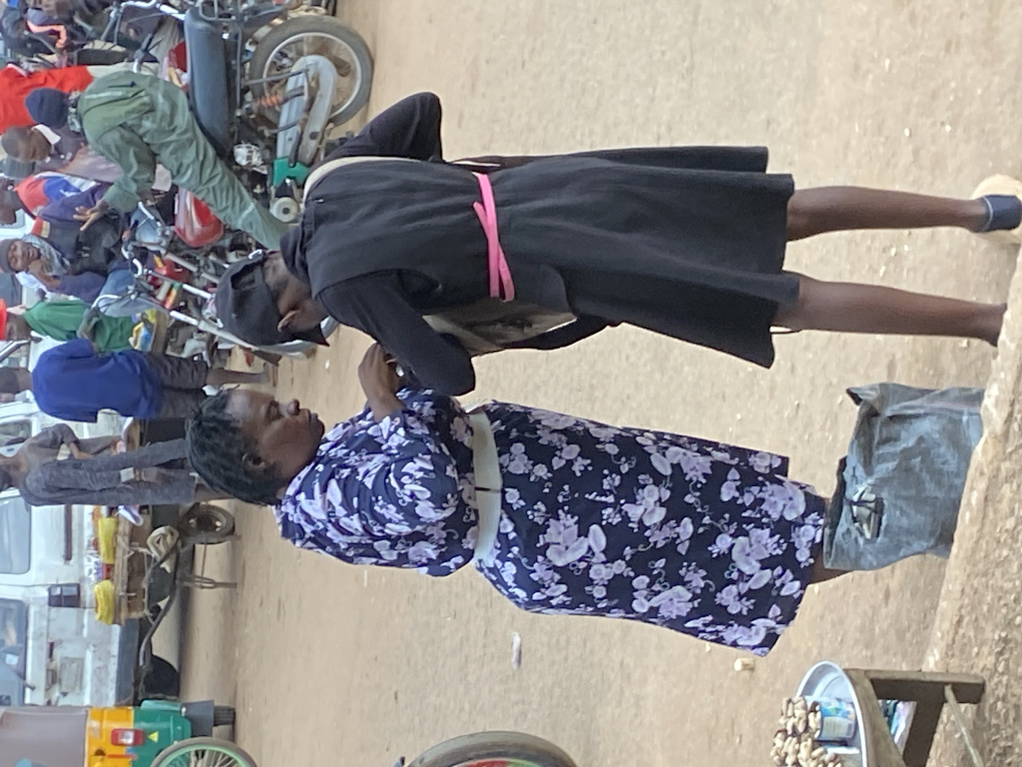 In Bukuru, Nigeria, a street market scene unfolds. Two women are the primary subjects. The woman on the left wears a dark blue floral dress with a white belt. She appears to be interacting with the woman on the right, who is dressed in a black outfit with a distinctive bright pink sash. The woman on the right is wearing a black cap and vest over a black dress. Their attention is focused on each other, possibly engaged in a transaction or conversation.
Behind them, the environment suggests a busy marketplace. A tricycle (Keke Napep) is visible to the left, a common mode of transport in Nigeria. Other individuals, mostly men, are seen in the background, some seated on motorcycles, indicating a bustling hub of activity. The ground is dirt, typical of many Nigerian streets. A cart with yellow goods is also present, likely for sale. A large, dark grey bag lies on the ground near the women. A small table with a tray of small food items is positioned in the foreground to the left. The overall atmosphere is one of everyday commerce and street life in an urban Nigerian setting.