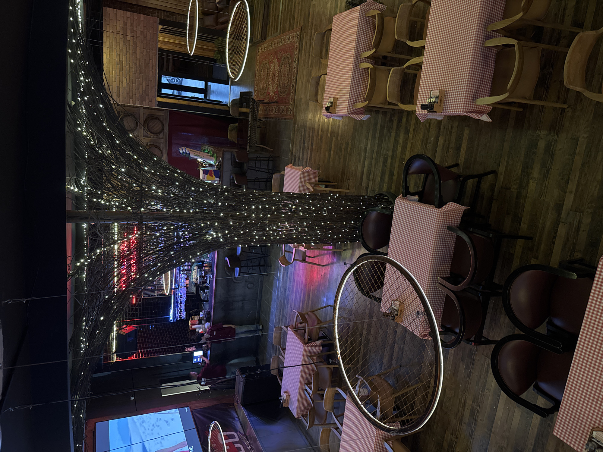 The image depicts a restaurant in Tashkent, Uzbekistan. The interior features a prominent decorative element resembling a tree made of branches and covered in numerous small lights. The dining area consists of tables covered with red and white checkered tablecloths, surrounded by various chairs, including wooden and upholstered designs. Circular light fixtures hang from the ceiling. A bar area is visible in the background, illuminated with neon lights and featuring bar staff. A large screen displaying content is also present in the foreground. The overall atmosphere is dimly lit and appears to be a combination of rustic and modern styles.