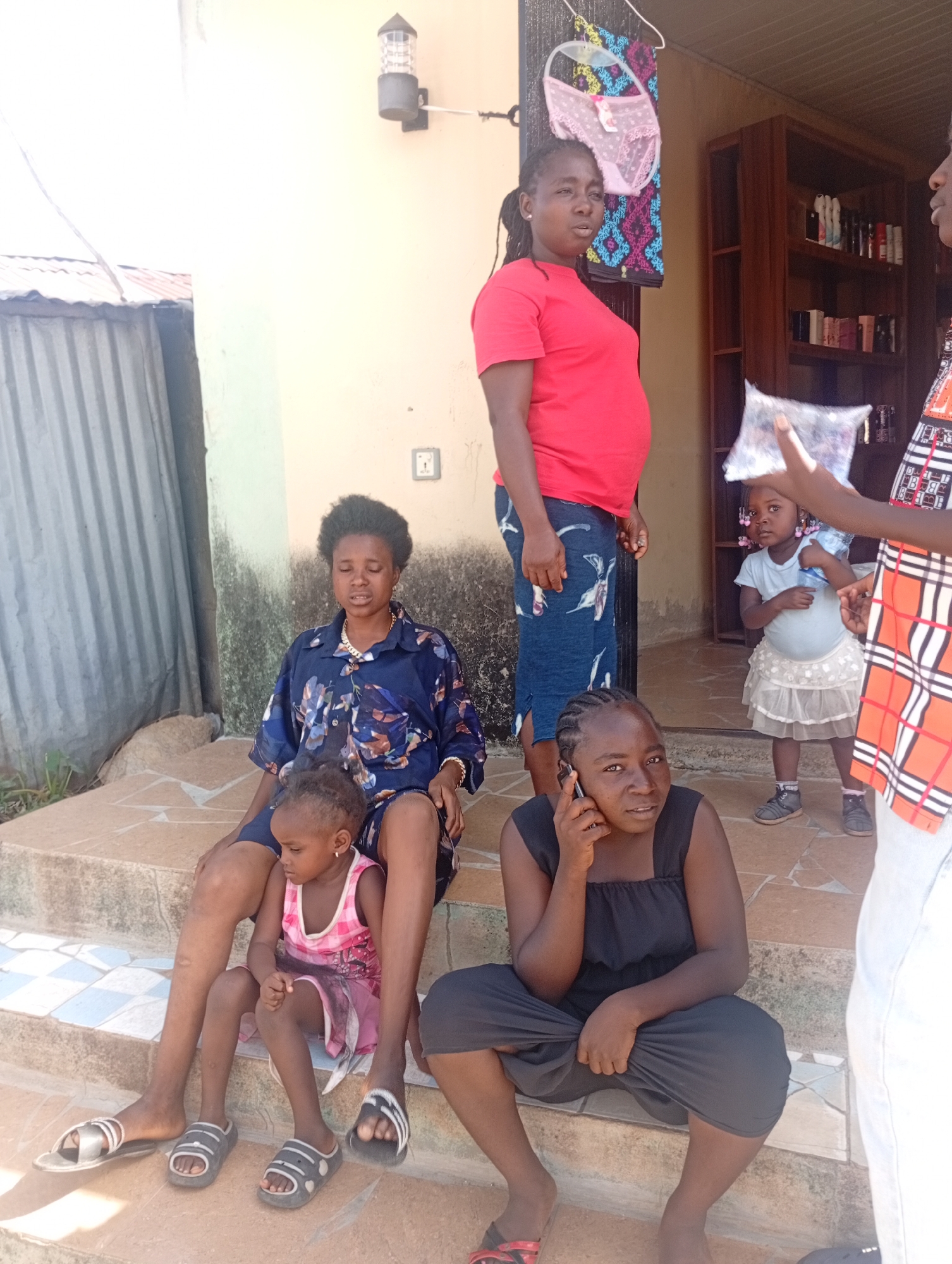 The image shows a group of people outside what appears to be a building in Jos, Nigeria. Four women and two young children are visible. One woman in a red t-shirt and patterned blue skirt stands near the entrance. Next to her is another woman, who appears to be handing her something in a plastic bag. There is a young girl in a light blue dress standing in front of them. Another woman sits on the steps with a young child in a pink dress on her lap. To the right is a woman talking on a mobile phone. A man in a patterned shirt is partially visible on the right edge of the frame. The surroundings include a building with an outdoor light, a metal structure on the left, and a shelf filled with items inside the building on the right. A garment hangs on the building next to the woman in the red shirt.