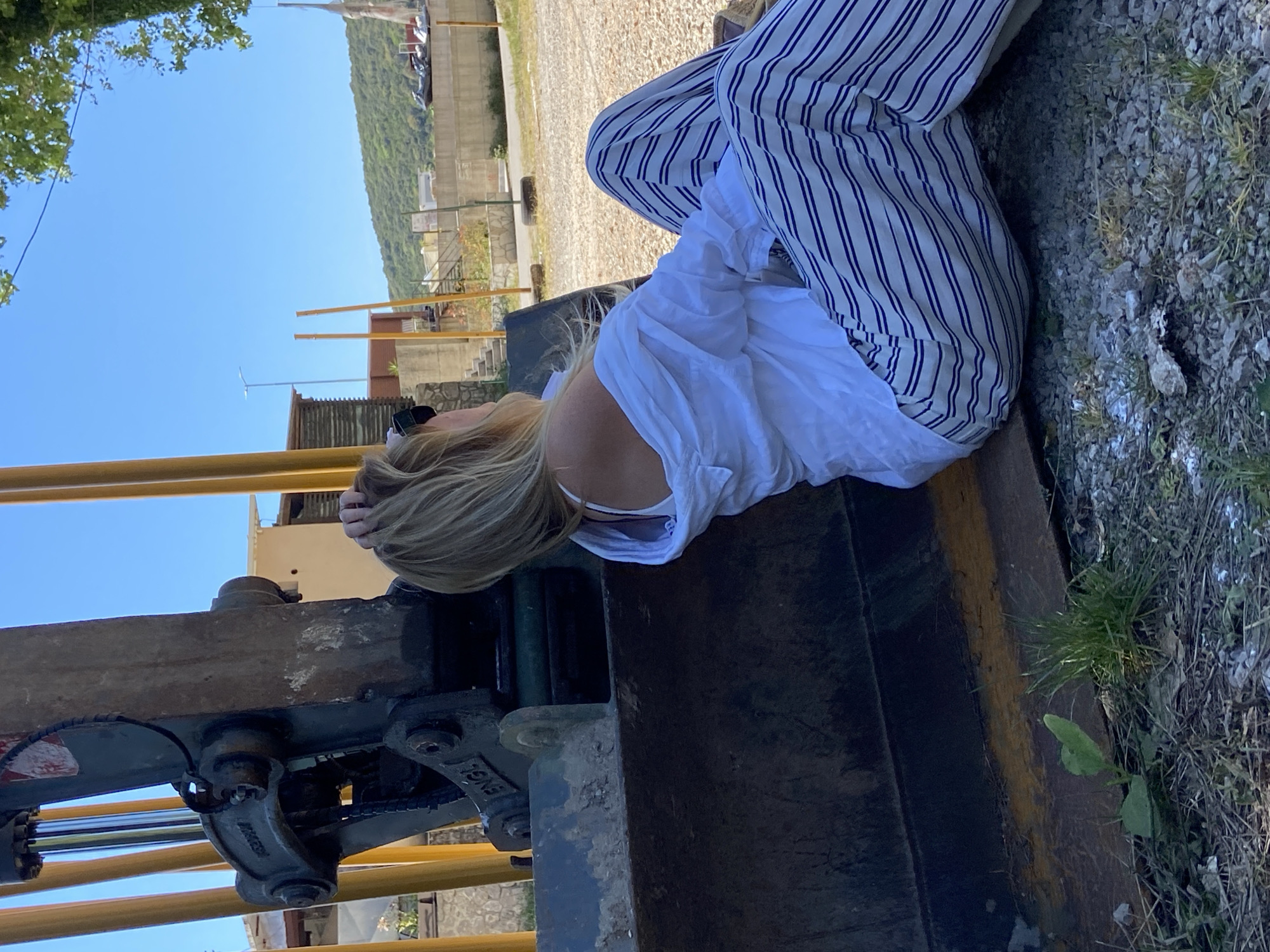 A woman with blonde hair is reclining on an excavator in Prijevor, Montenegro. She is wearing striped pants and a white shirt. The excavator is positioned in a gravelly area with sparse vegetation. In the background, there is a building and a mountain. The sky is clear and blue.
