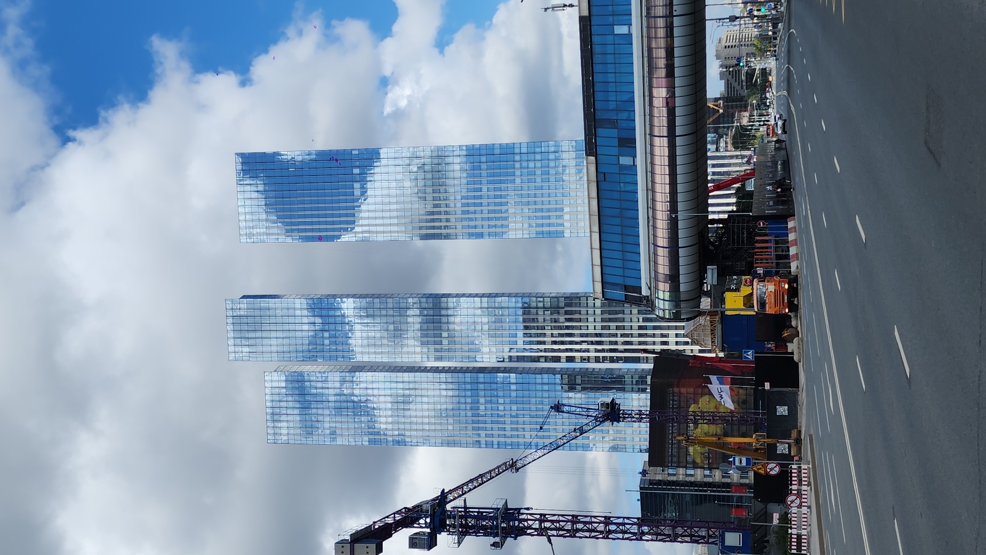 The image captures a contemporary urban landscape in Moscow, Russia, likely near the Moscow City business district, characterized by modern skyscrapers and ongoing construction. In the foreground, a multi-lane asphalt road stretches into the distance, marked with white lines. To the left, a tall blue construction crane dominates the scene, with its lattice boom extending diagonally. Adjacent to the crane, several buildings are visible, some under construction, with fencing and construction materials. An orange truck is parked on the right side of the road, near the construction site. A prominent feature is the elevated glass walkway that spans across the road, connecting buildings and offering a clear view of the sky. In the background, three towering skyscrapers with reflective glass facades stand tall, their surfaces mirroring the cloudy blue sky. The sky itself is a mix of dramatic dark and light clouds against a vibrant blue backdrop. A few small purple objects, possibly kites or balloons, are visible against the sky. The overall impression is one of dynamic urban development and modern architecture.