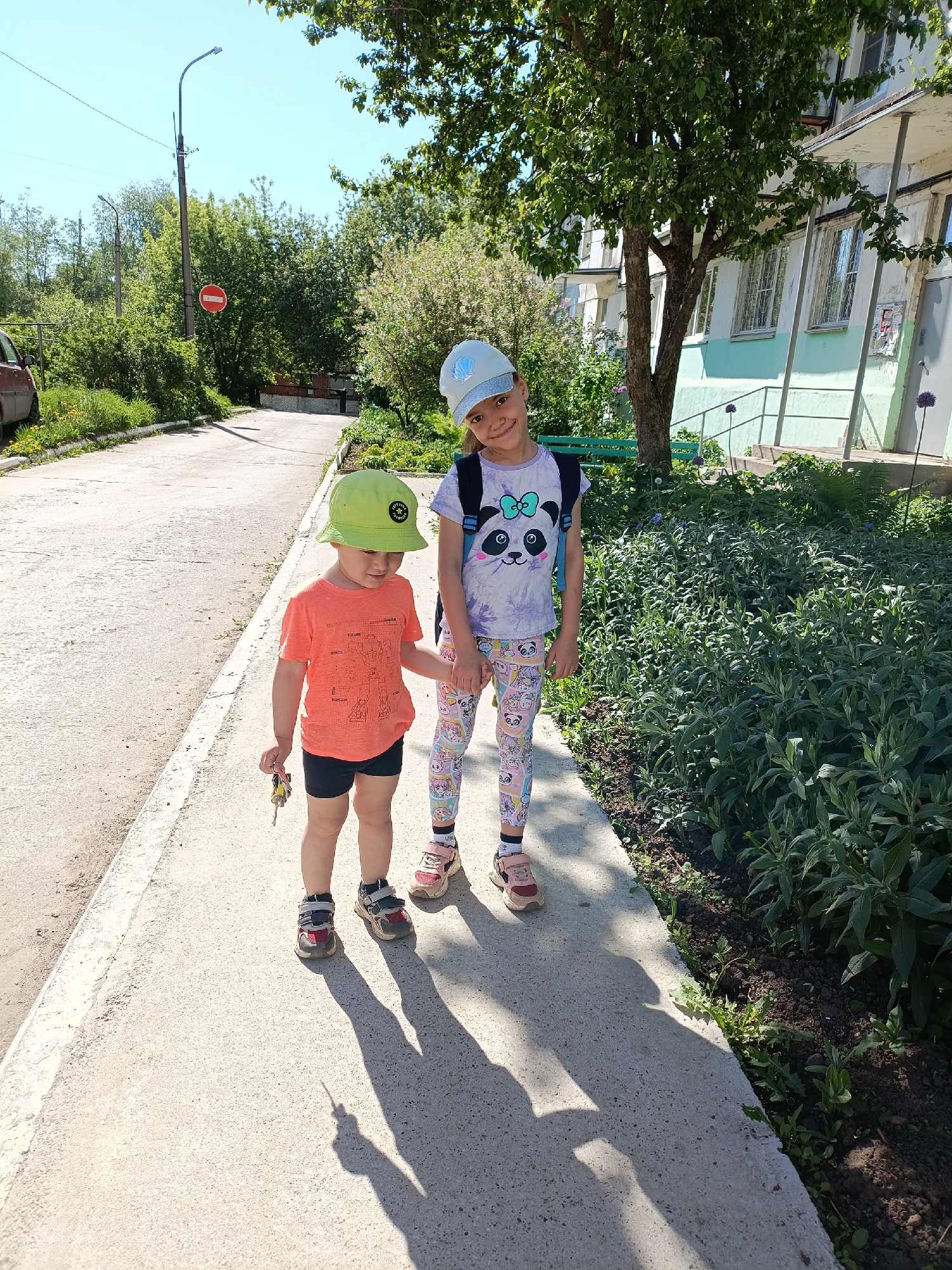 Two children are standing on a sidewalk adjacent to a building in Chaikovsky, Russia. The older child, on the right, wears a light blue cap, a panda-themed t-shirt, leggings with cartoon characters, pink and brown sneakers, and a light blue backpack. She is standing and slightly smiling at the camera. The younger child, on the left, wears a lime green bucket hat, an orange t-shirt with a robot-themed design, black shorts, and red and gray sneakers. He is holding a small, possibly toy-related, item in his left hand. Both children are holding hands. The sidewalk is concrete and appears to be in an urban or suburban setting. A garden bed is alongside the sidewalk, filled with green foliage and some blooming purple flowers. In the background, there is a building with light green and white walls and visible windows. Beyond the building, various trees and greenery fill the scene. A road with a "no entry" traffic sign is also visible further in the background to the left. The scene is bright, with shadows suggesting it is daytime and sunny.