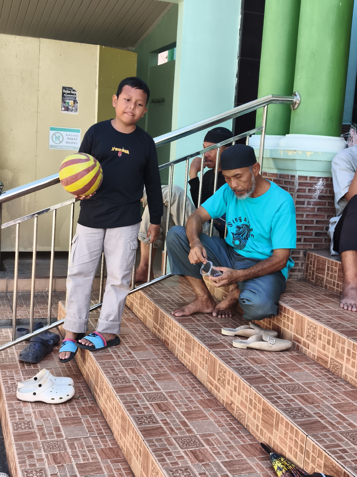 In Tonasa, Indonesia, a boy in a black long-sleeved shirt with a Spider-Man logo and light-colored cargo pants stands on outdoor steps, holding a yellow and red striped ball. He is facing forward and appears to be looking at the camera. To his right, an older man wearing a blue t-shirt and dark pants sits on the steps, fiddling with a small clear object in his hand. Several other people are visible in the background, also seated on the steps. Various footwear, including sandals and white Crocs, are scattered on the steps. The environment appears to be the exterior of a building with a light green wall and green pillars. A metal railing runs along the steps. The steps are tiled with a brown and beige patterned ceramic. There are also signs on a wall behind the boy, one with text in Indonesian.