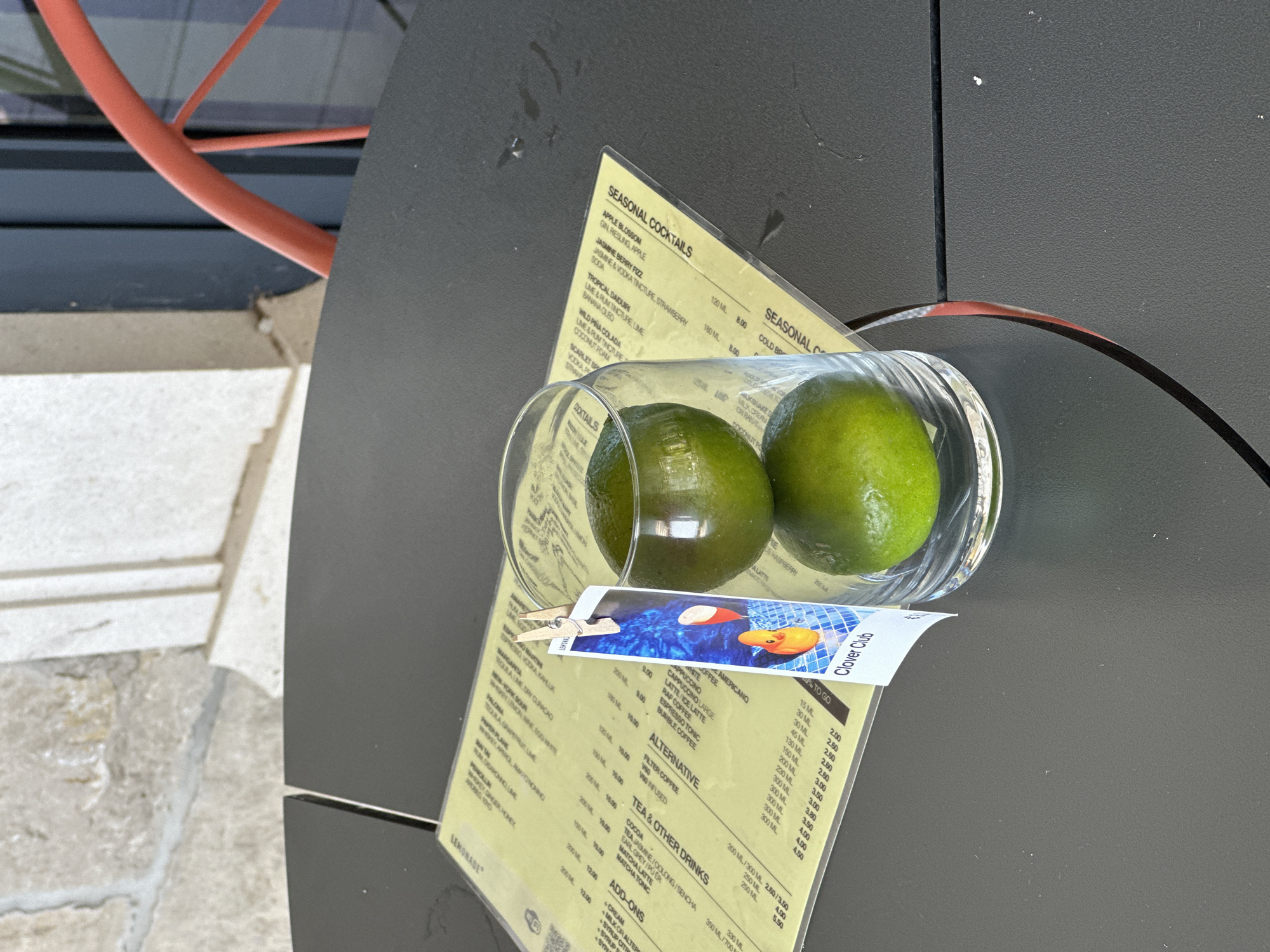 On a dark gray outdoor table in Rafailovići, Montenegro, a clear glass contains two green limes stacked on top of each other. A menu, primarily in yellow, is spread out on the table, partially obscured by the glass. A small card, attached to the glass with a wooden clip, displays a cartoon duck and the text "Clover Club." The background shows a light-colored stone wall and a dark blue structure. A portion of a red chair is visible on the right side of the frame.