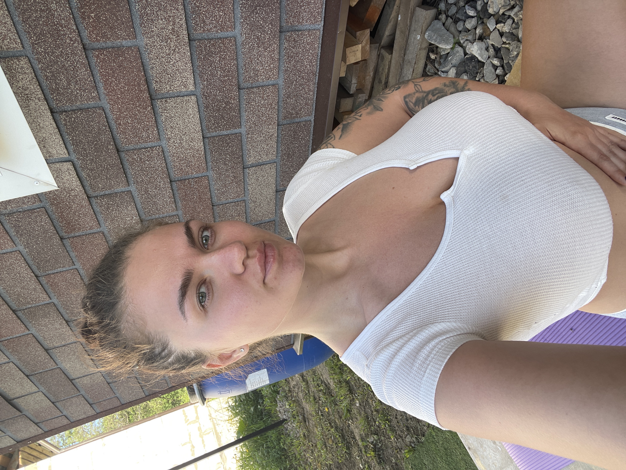 A woman with light skin, light green eyes, and dark eyebrows is the primary subject of this eye-level shot. She has a nose piercing and her hair is pulled back. She is wearing a white short-sleeved shirt. Part of a tattoo is visible on her left arm.
The background environment appears to be outdoors. A brick structure is to the upper left, with stacked wood behind her. The ground consists of grass, vegetation, and gravel. A blue container is partially visible. In the distance is a wire fence with greenery.
The exact location within Мосметаллоконструкция Онт, Russia is not discernible from the available visual data.