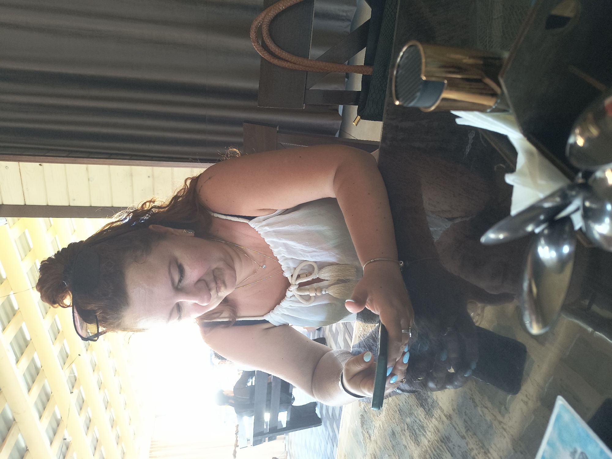 A middle-aged Caucasian woman with auburn hair pulled into a bun, wearing a white camisole, is seated at a table outdoors, looking down at her smartphone. She has light skin, with rouged cheeks and defined eyebrows, and she is wearing sunglasses pushed up on her head. She has light blue nail polish on her fingernails and is wearing silver bracelets on both wrists. The table she is sitting at appears to be made of a dark, possibly glass, material with a marbled pattern. In front of her, on the table, are a white napkin dispenser, a gold and black object that appears to be a salt or pepper shaker, and some silverware. In the background, to the left, there are other people seated at a dark wooden bench, and the area is covered by a light-colored, slatted roof structure. To the right of the woman, a dark wooden chair is visible, and a woven handbag with tan straps rests on the chair. The overall setting suggests an outdoor seating area of a restaurant or cafe in Solotvyno, Ukraine.