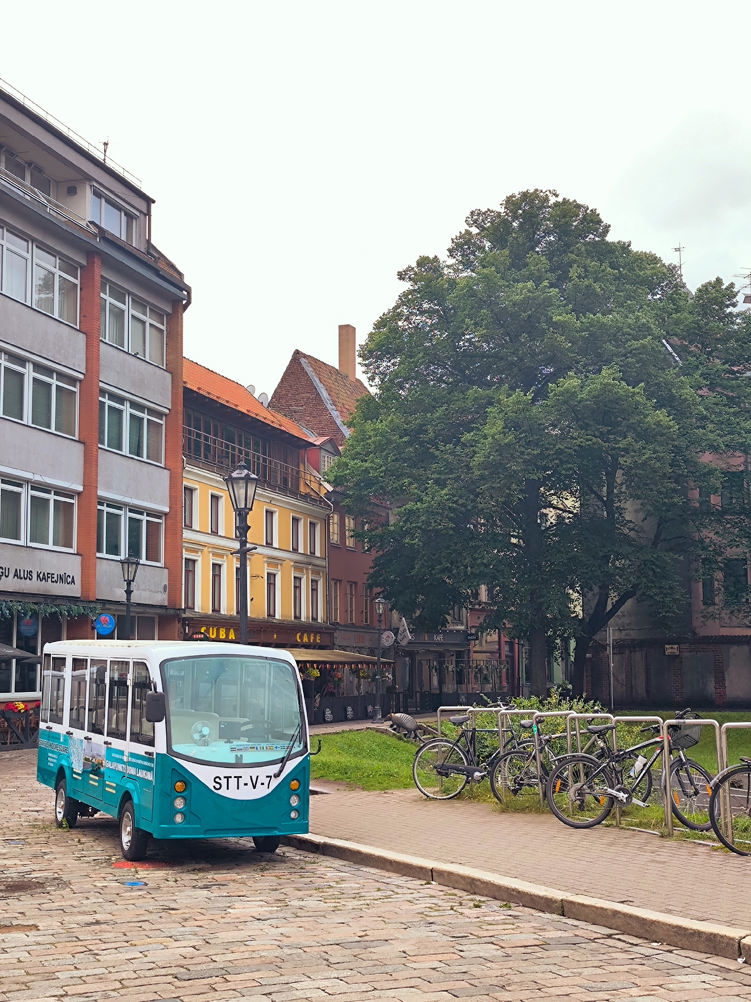 A teal and white tourist shuttle bus, license plate STT-V-7, is parked on a cobblestone street in Riga, Latvia. The bus is positioned on the left side of the frame, facing slightly to the right. In the background, a row of buildings with orange tiled roofs line the street, featuring shops and cafes, one labeled "CUBA CAFE". A large, mature tree with dense green foliage dominates the right side of the image. To the right of the tree, more buildings are visible, including a brick structure. A row of bicycles is parked in front of a metal rack on the sidewalk, separating the street from a grassy area. The sky is overcast.