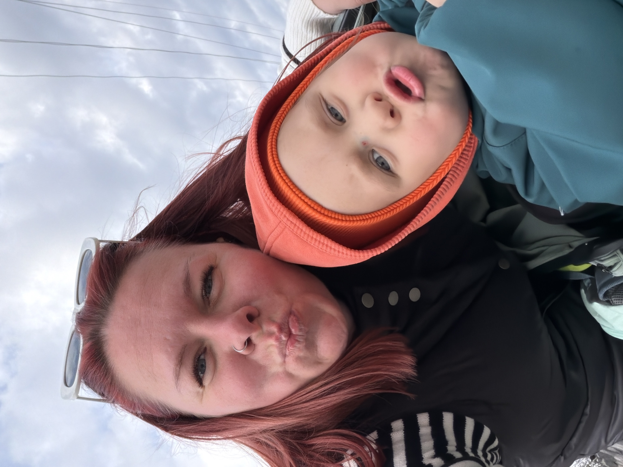 The image, oriented on its side, captures a close-up selfie of two individuals: an adult woman and a young child, likely outdoors under an overcast sky.
The woman, positioned in the foreground and to the left when rotated correctly, has reddish-brown hair and fair skin. She wears sunglasses pushed up on her head and has a nose piercing. Her lips are puckered in a playful "duck face" expression, and she has visible facial details including rosy cheeks. She is dressed in a black jacket with silver buttons and a black and white striped garment partially visible.
The child, positioned above and to the right of the woman, also makes a similar puckered lip expression. The child has fair skin and light-colored eyes, wearing an orange hooded garment underneath a blue or teal outer jacket.
The background consists of a cloudy, light gray sky with some wispy cloud formations. Thin power lines are visible stretching across the upper portion of the sky, indicating an outdoor setting. The overall mood appears lighthearted and playful, capturing a shared moment between the two subjects. No discernible text is visible in the image.
