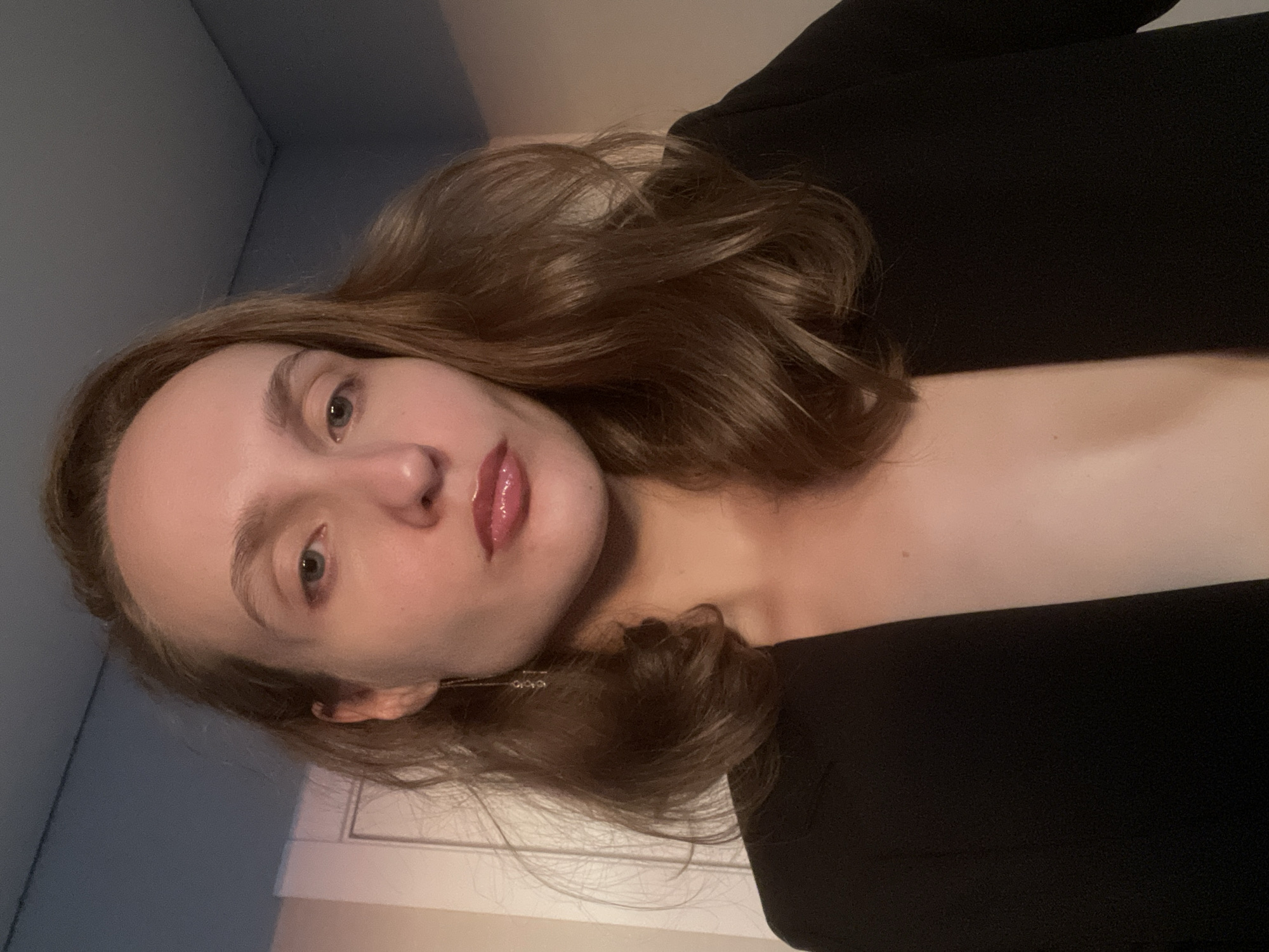 A person with light skin, medium-brown hair, and blue eyes is looking forward at the camera. They are wearing a black blazer with a low-cut neckline, revealing their chest. The background is blurred, with what appears to be a wall and a door visible. There is no indication of the location being in the city of Подшивалово, Russia.