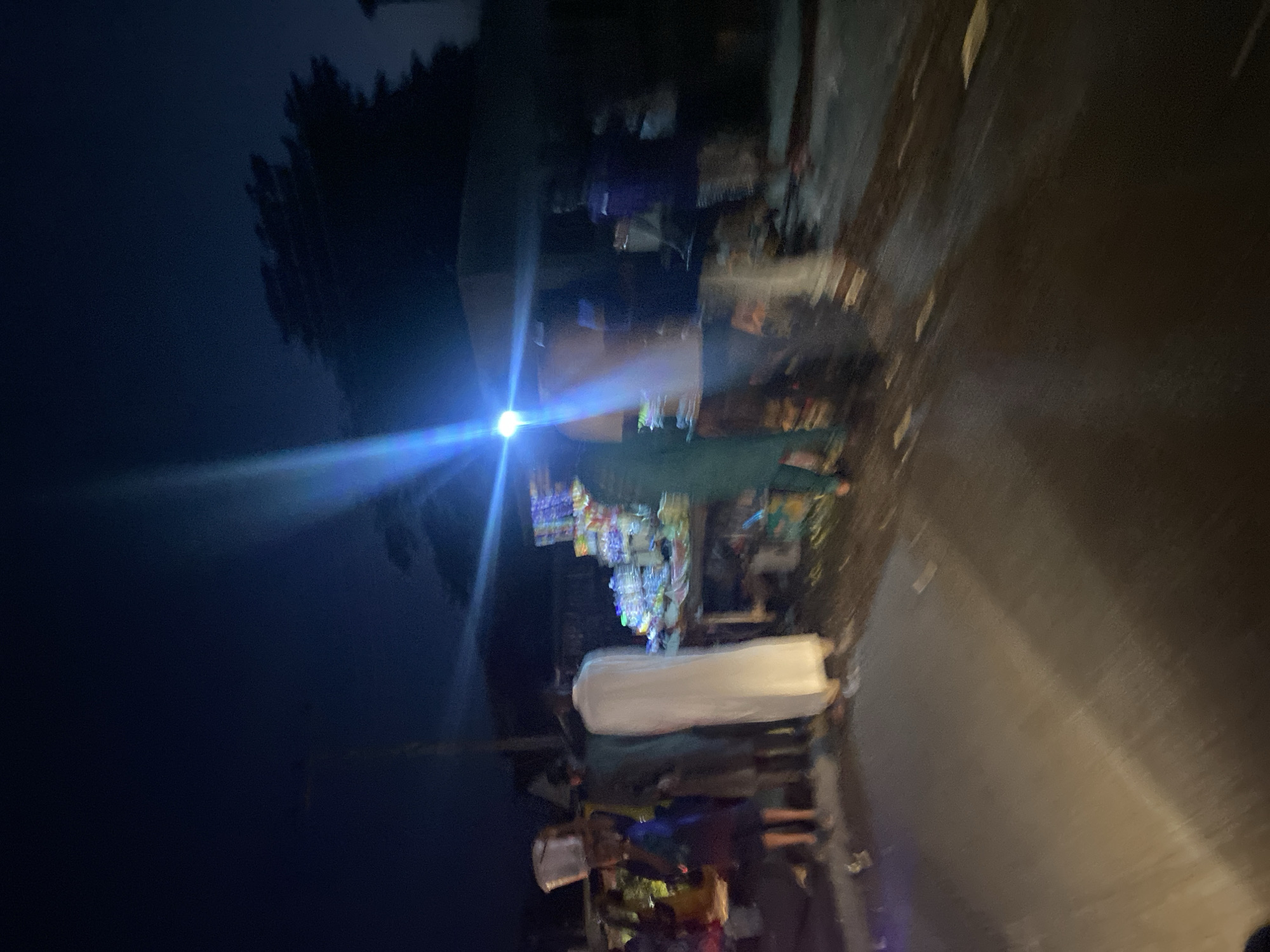 The image captures a nighttime scene at a roadside market in Jalingo, Nigeria. A bright blue light, likely from a streetlamp or vehicle, casts a strong beam across the frame, creating lens flares and obscuring some details. Several people are visible, most of whom are blurred due to motion or low light conditions.
In the foreground, a dark, wet road surface is visible, with reflections of light. To the left and center, a group of individuals stand around a market stall laden with goods, including what appear to be bottled drinks and packaged items. One person in the center is wearing a long, white garment. Further left, another person carries a white bucket on their head.
To the right of the stall, more people are present, some appearing to be customers and others vendors. The environment suggests an open-air market operating after dark, with temporary structures and goods displayed on tables and possibly on the ground. The overall impression is one of a bustling, if dimly lit, commercial activity at night.