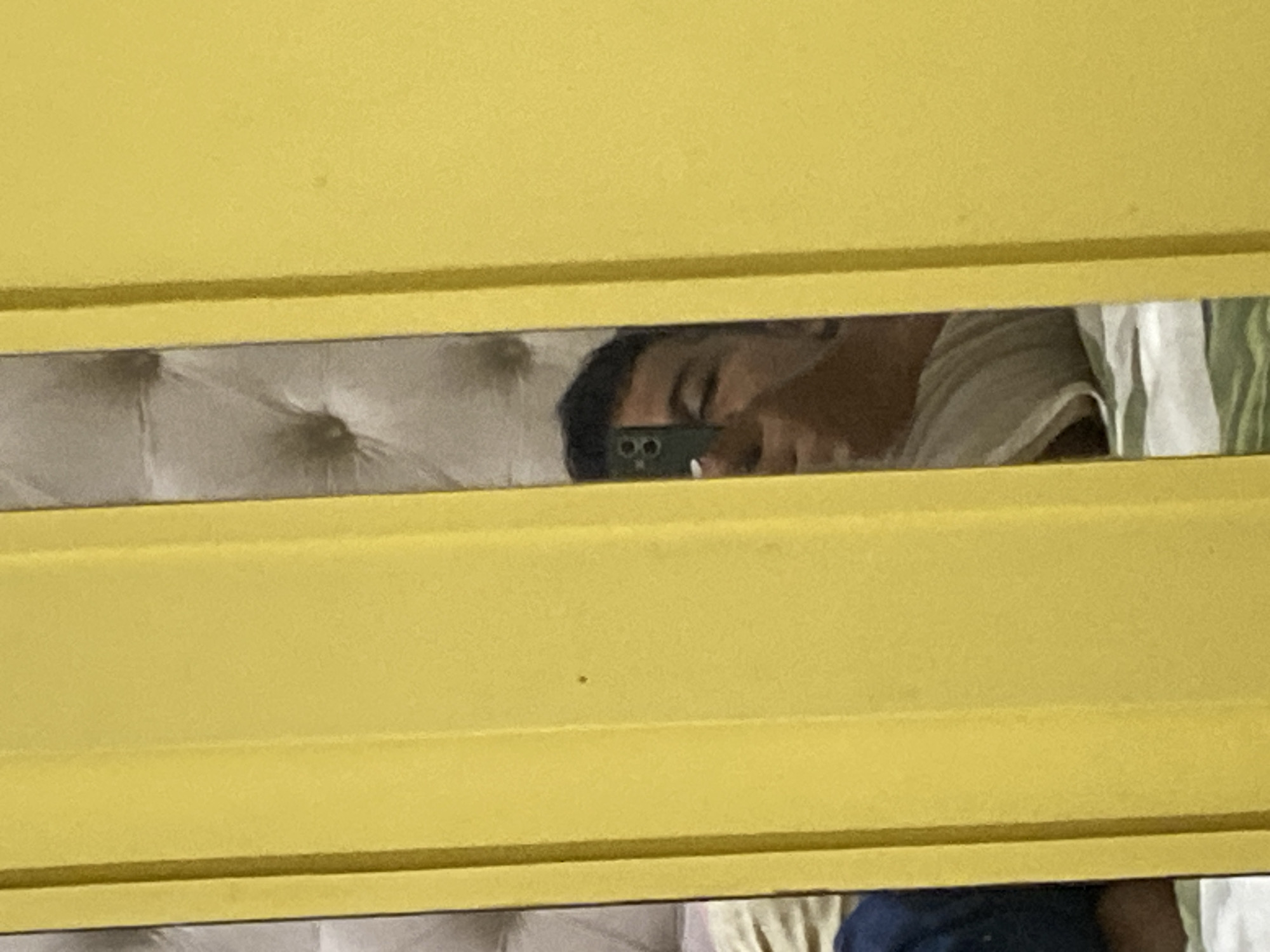 In a bedroom in Lambaro Kueh, Indonesia, a person is reflected in a mirror, taking a selfie with a smartphone. The mirror is positioned between two yellow panels, which appear to be part of a closet or wardrobe. The person is holding a dark-colored smartphone horizontally, aiming it towards their face. The reflection shows the person's face, with one eye visible and the other partially closed or squinting. Their hair is dark and cut short. The background behind the person in the reflection is out of focus but appears to be a tufted headboard. The overall lighting suggests an indoor setting, possibly during the day.