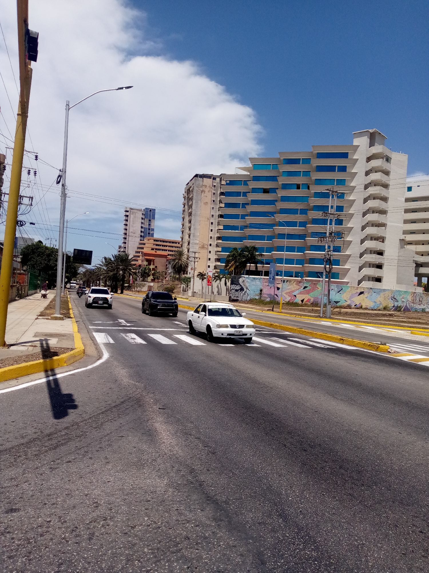 The image depicts a street scene in Porlamar, Venezuela, characterized by a wide, multi-lane road with crosswalks. The road is primarily gray asphalt, with yellow and white painted curbs and lane markings. Three vehicles are visible on the road: a white sedan, a black SUV, and a white SUV further in the distance.
The backdrop is dominated by tall buildings, notably a white building with a distinct stepped facade and blue-tinted windows. A mural adorns the wall along the street. Overhead utility lines and poles are visible. The sky is blue with scattered white clouds. The angle of the shadows suggest it is midday. There is a light traffic light on the far left of the image.