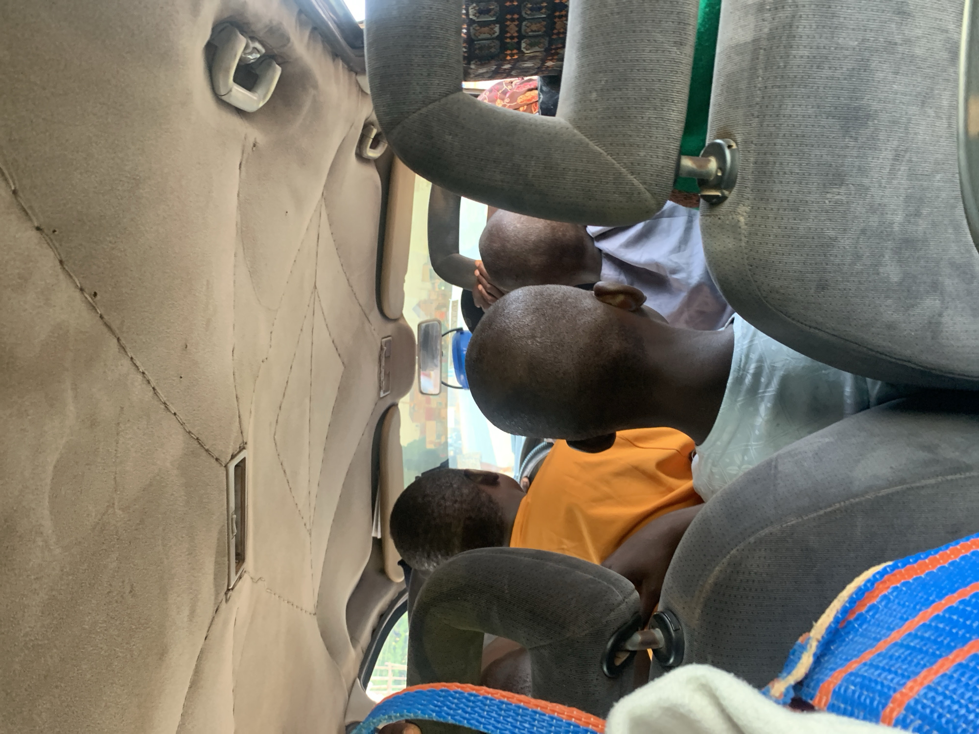 Inside a vehicle in Keffi, Nigeria, multiple people are seated. The interior features textured beige upholstery. Passengers' clothing varies; one wears an orange shirt, another a light-colored patterned shirt. The car seats are upholstered in gray fabric with green and blue accents.