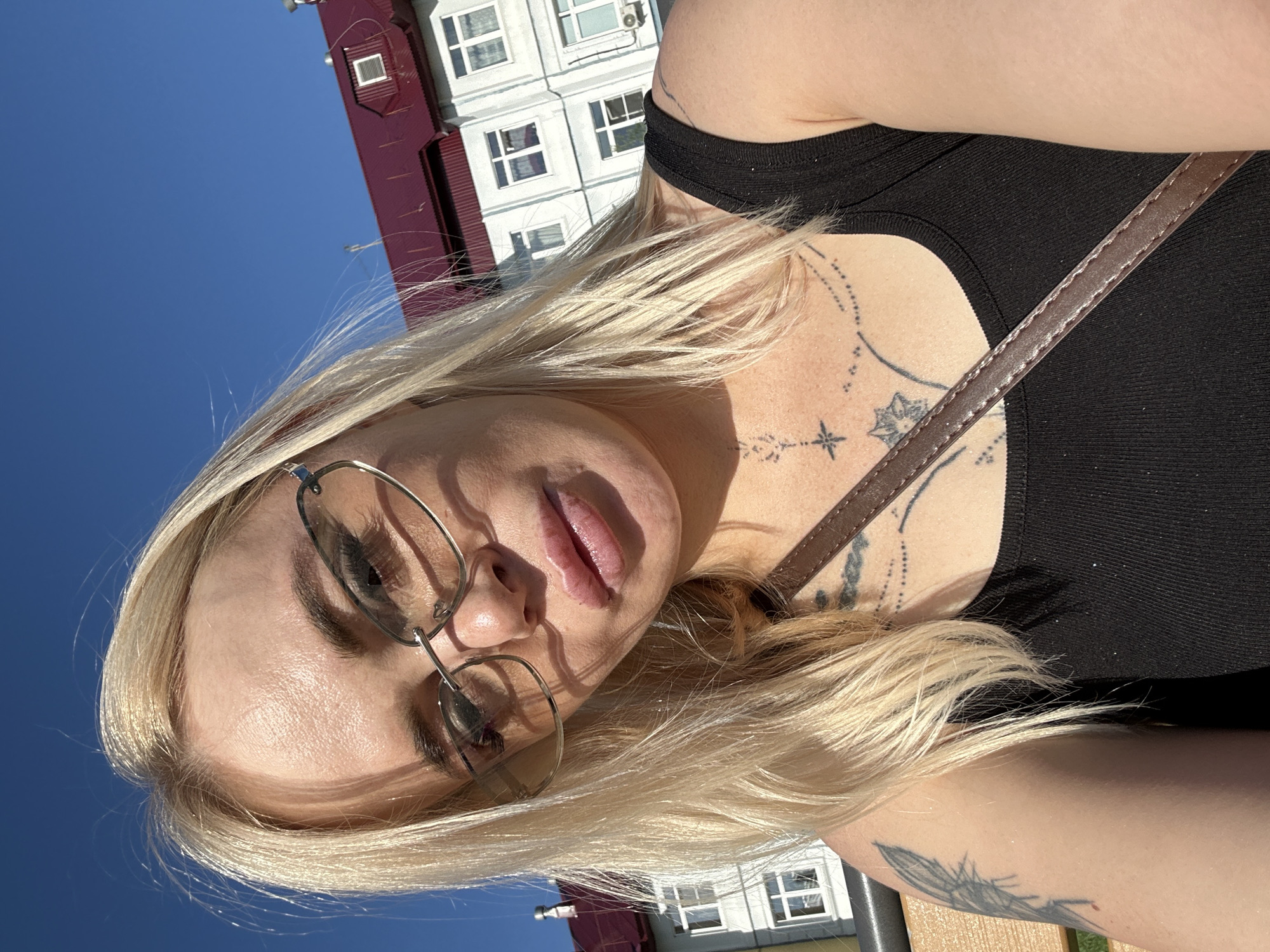 In a close-up, outdoor selfie, a blonde woman with light skin, wearing glasses and a black tank top, is pictured against a bright blue sky and a building with a red roof. Her face, partially obscured by her hair and glasses, shows her lips slightly parted and her eyes looking towards the camera. Visible tattoos adorn her chest and left arm. The building in the background has multiple windows and appears to be a multi-story structure. The lighting suggests it is daytime, with shadows cast across the woman's face and hair. The overall scene is a casual, sunlit portrait.