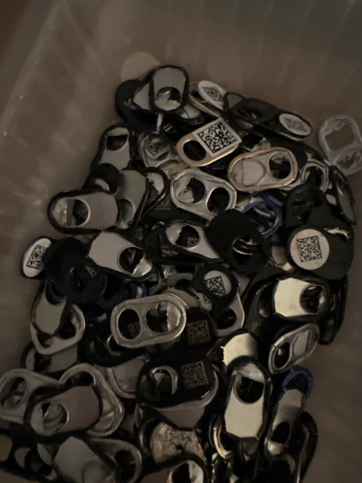 The image shows a pile of aluminum pull tabs, some with black plastic or painted coatings, and a few with QR codes printed on them. The tabs are piled loosely in what appears to be a clear plastic container or bag. The lighting is dim, creating shadows and making it difficult to discern fine details. The background is out of focus and appears to be a neutral, indoor environment. There is no information in the image to suggest a specific location in Nevinnomyssk, Russia.