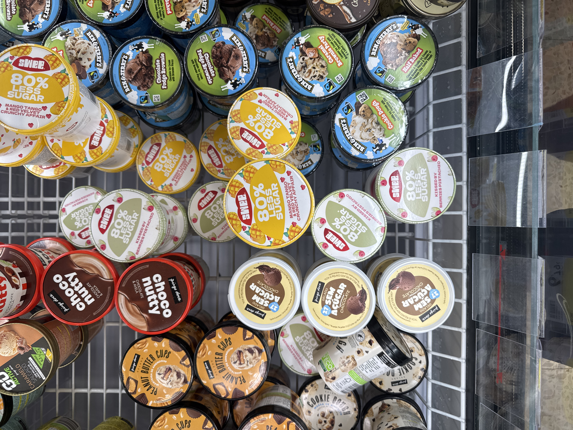 The image shows a close-up, overhead view of an ice cream display in a refrigerated case. The display is filled with various tubs of ice cream, primarily from brands like "Pingo Doce," "Swee," and "Ben & Jerry's." The ice cream tubs are arranged in rows and stacks, with some labels visible. Examples of visible flavors include "Choco Nutty," "Peanut Butter Cups," "Mango Tango & Red Velvet Crunchy Affair," "Kissed by Berries Pistachio," "Chocolate Fudge Brownie," "Caramel Brownie Party," and "Cookie Dough." The tubs are predominantly cylindrical with colorful labels. The environment appears to be a retail setting, likely a supermarket or grocery store, with the metallic shelving of the refrigerated unit visible beneath the ice cream. The lighting is even, and the image is sharp, allowing for clear identification of the products. There are no people or discernible actions depicted in the image. The image was captured in a commercial freezer in a grocery store located in the city of Castelo Branco, Portugal.