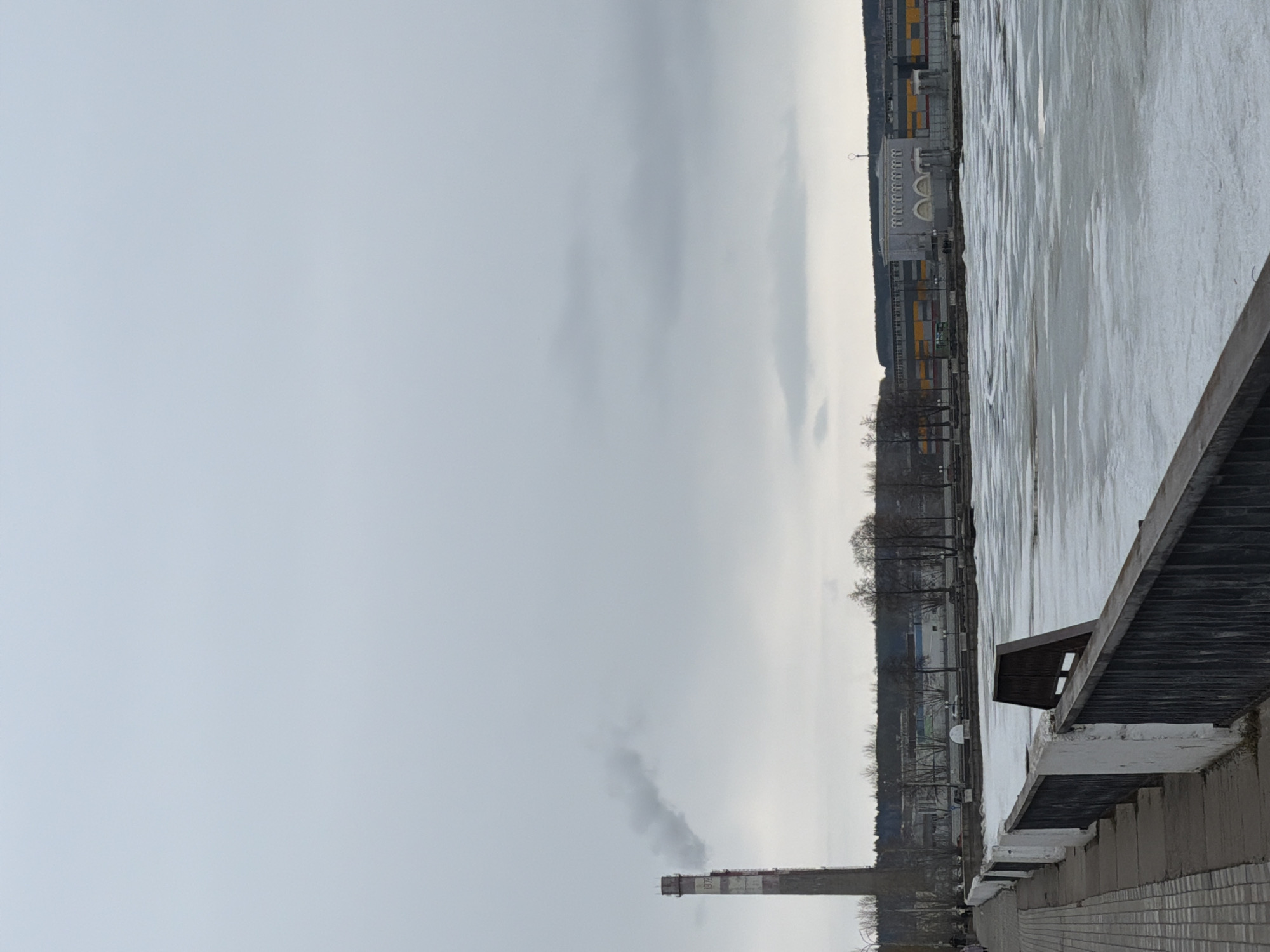 The image depicts a view of the industrial landscape in Votkinsk, Russia, characterized by a somber, overcast sky and a partly frozen body of water. A tall smokestack emits a plume of smoke, indicating industrial activity. The near foreground features a concrete embankment with wooden structures or fencing. The body of water, potentially a river or lake, is partially covered with ice, suggesting cold weather conditions. Across the water, industrial buildings and trees line the bank, with the architecture reflecting the area's industrial heritage. The lack of vibrant colors and the overcast sky create a muted and bleak atmosphere.