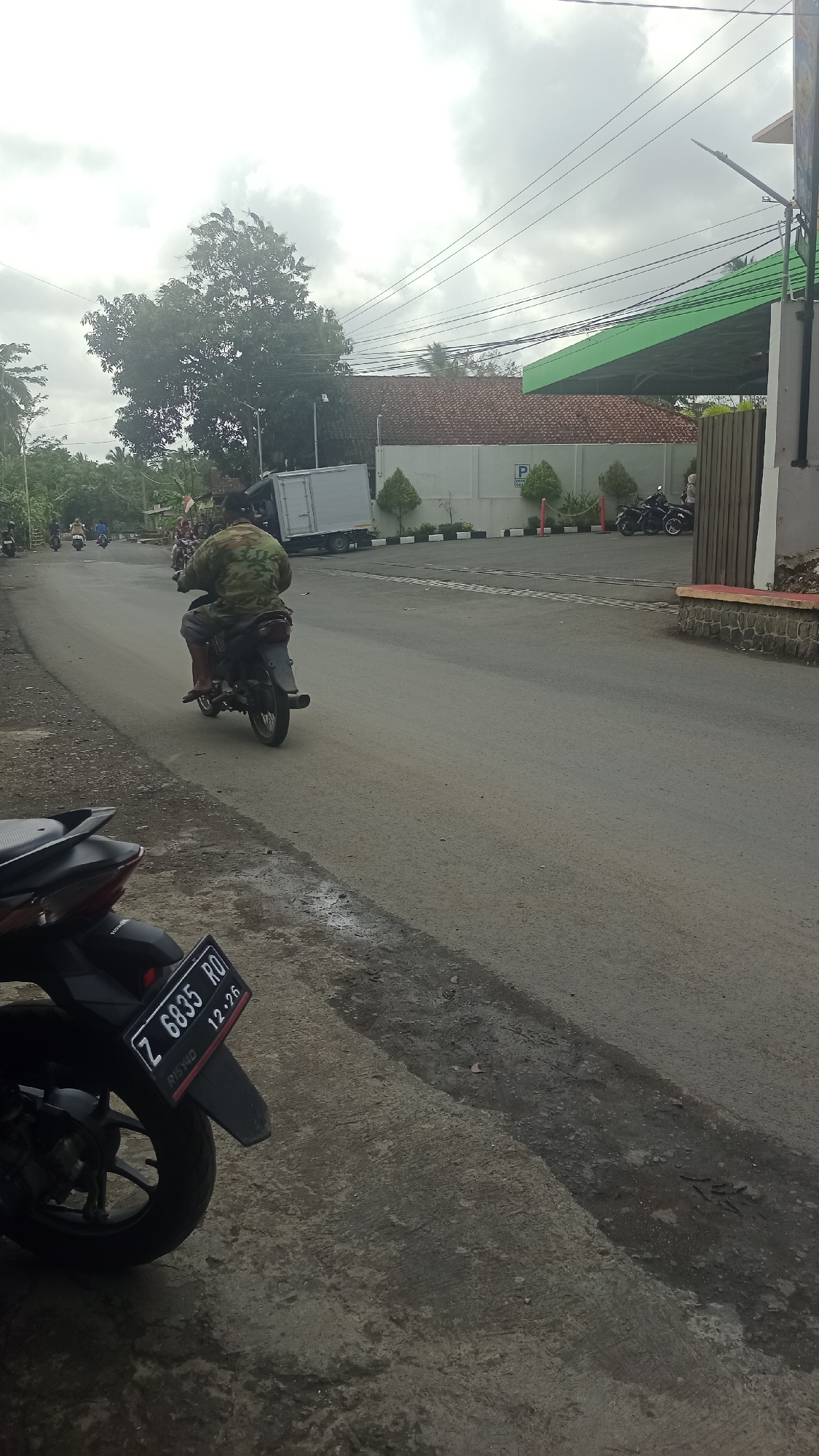 A man wearing a camouflage jacket rides a motorcycle on a paved road in Cikalong, Indonesia. The man is viewed from behind, and his face is not visible. The motorcycle is dark-colored, and the man's legs are bare. The road is wet and appears to have recently rained. In the background, a light-colored box truck is parked on the side of the road, and a building with a red tiled roof is visible. There are trees and power lines in the distance. A black motorcycle with a license plate reading "Z 6835 RO" is parked in the foreground, facing to the left. The sky is overcast and cloudy.