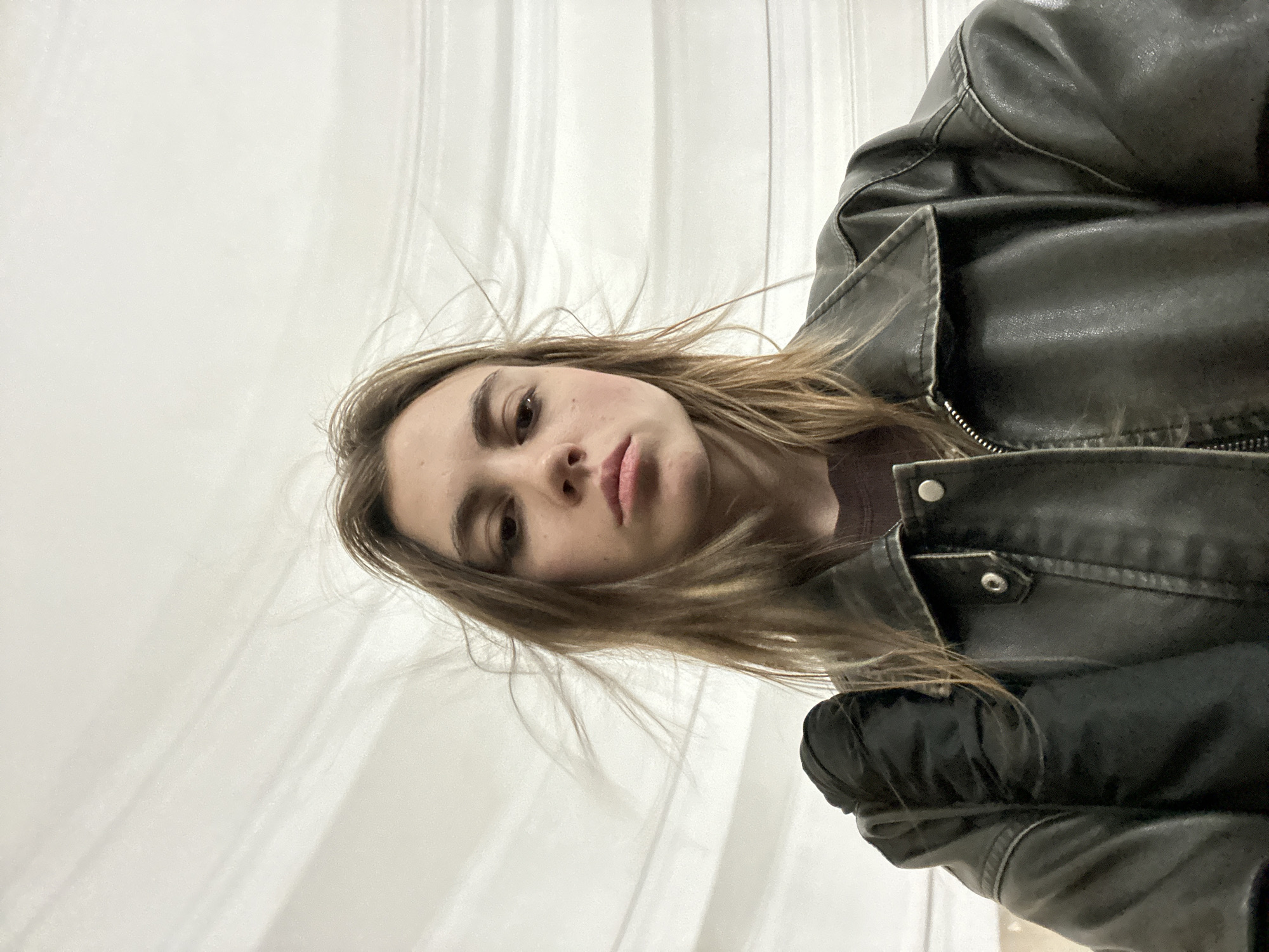 A young woman with shoulder-length brown hair, fair skin, and light eyes is captured in a selfie from a low-angle perspective. She is wearing a dark-colored leather jacket over a brown t-shirt. Her hair is windswept, suggesting an outdoor or drafty environment. The background consists of curved, light-colored surfaces with horizontal lines, possibly indicative of a modern architectural structure. The lighting is even, illuminating her face and the textures of her clothing. The setting is likely an urban environment, given the style of the jacket and the backdrop. The specific location within Moscow, Russia, cannot be determined from the visual information provided.