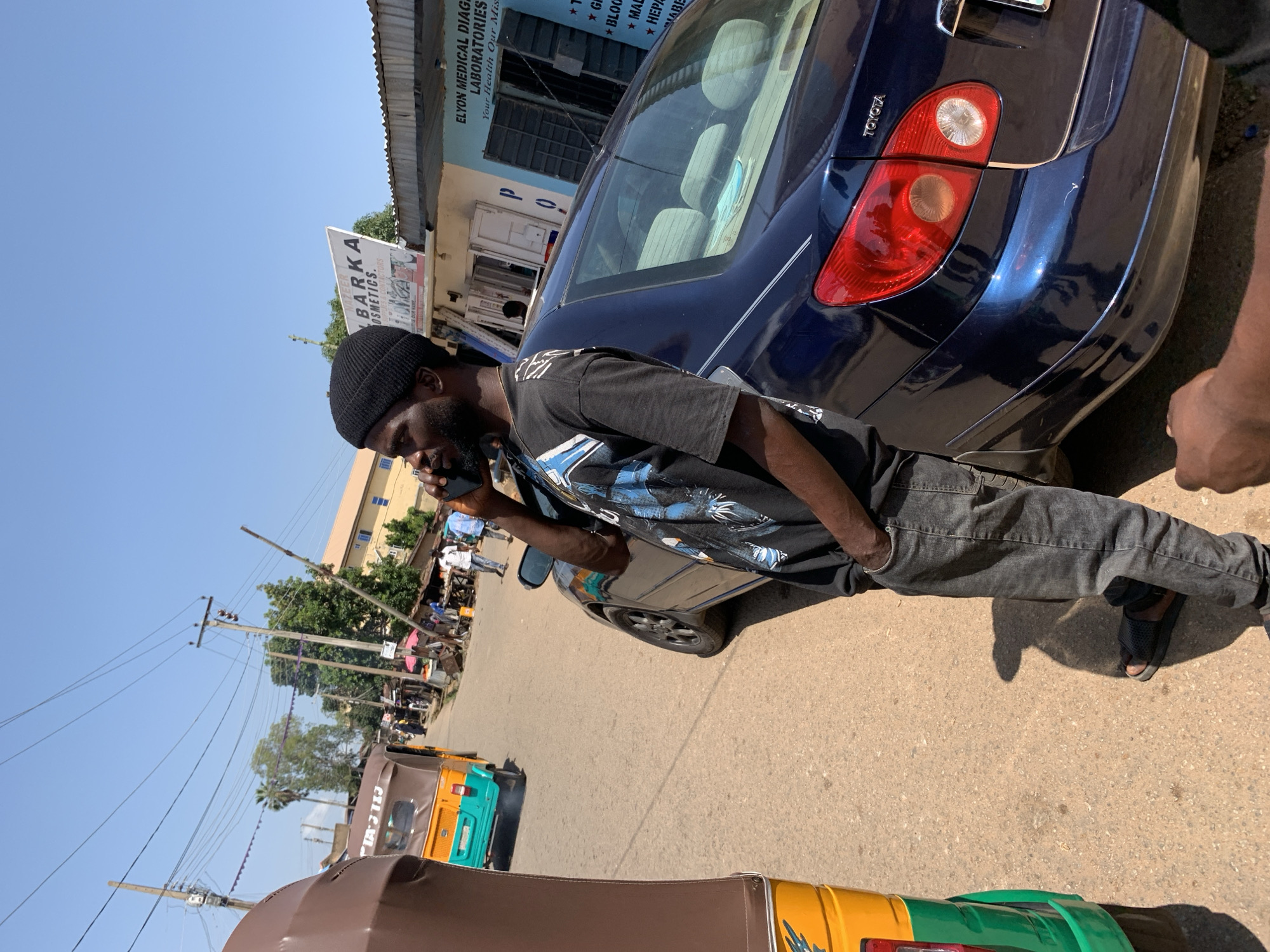 A Black man, wearing a black beanie, black t-shirt with a blue graphic, and grey jeans, stands on a paved street in Jos, Nigeria, holding a mobile phone to his ear. He is standing next to a dark blue Toyota sedan. To his left, a portion of a brown and yellow auto-rickshaw is visible. In the background, there are buildings, other vehicles, people walking, and overhead power lines. The sky is clear and blue.