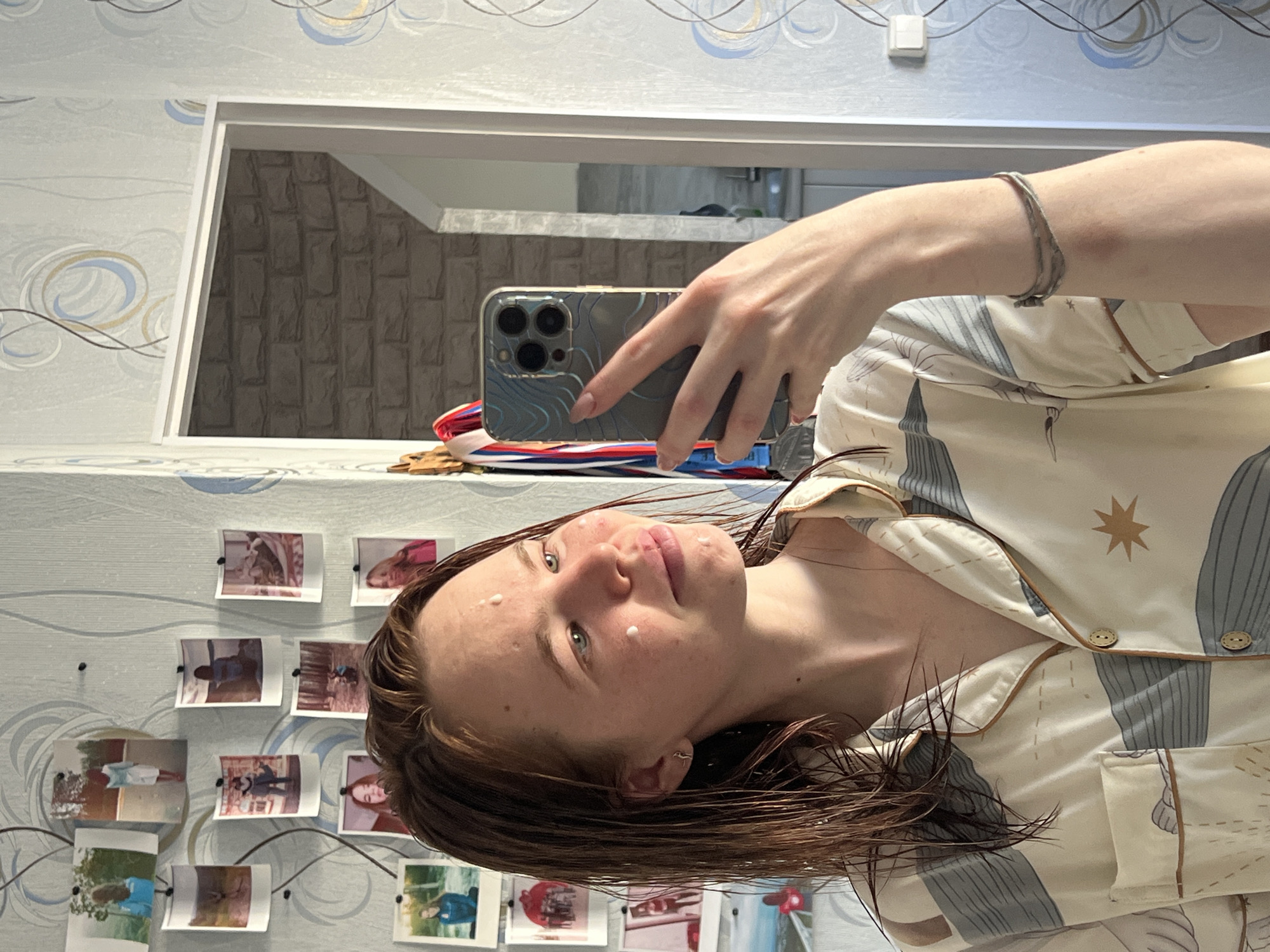 A woman takes a selfie in front of a mirror. She holds an iPhone 13 Pro in a gray topographical patterned case. The woman has wet brown hair and is wearing a patterned pajama shirt. There are small dots of cream on her face. A silver bracelet is visible on her wrist. Behind the phone are medals and ribbons on a surface that appears to be a shelf. The mirror reflects a gray brick wall. To the left of the mirror, the wall is decorated with photographs pinned to the wall. The wallpaper has a swirling pattern of blues and browns. The location is Chaikovsky, Russia.