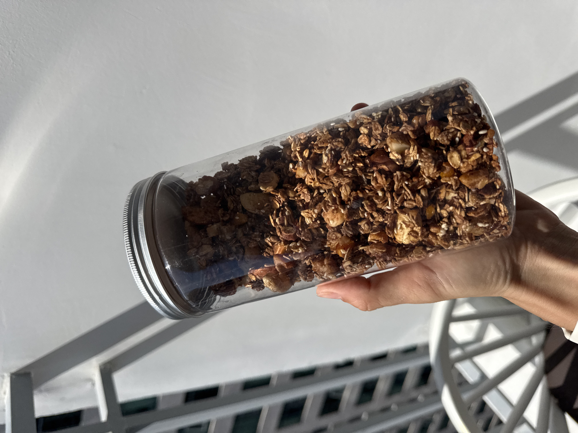 A hand holds a clear plastic jar filled with granola, which is positioned in the foreground against a bright, white background. The granola appears to be a mix of oats, nuts, and seeds, with a rich brown color. Behind the jar, out of focus, is a white railing, possibly part of a balcony or fence, casting a shadow on the wall. To the left of the railing, a blurred image of a modern building's facade with dark rectangular windows is visible, suggesting an urban setting. The lighting indicates it is daytime, likely outdoors, with strong sunlight creating distinct shadows. The location is identified as Bac Nha Trang Ward, Vietnam.