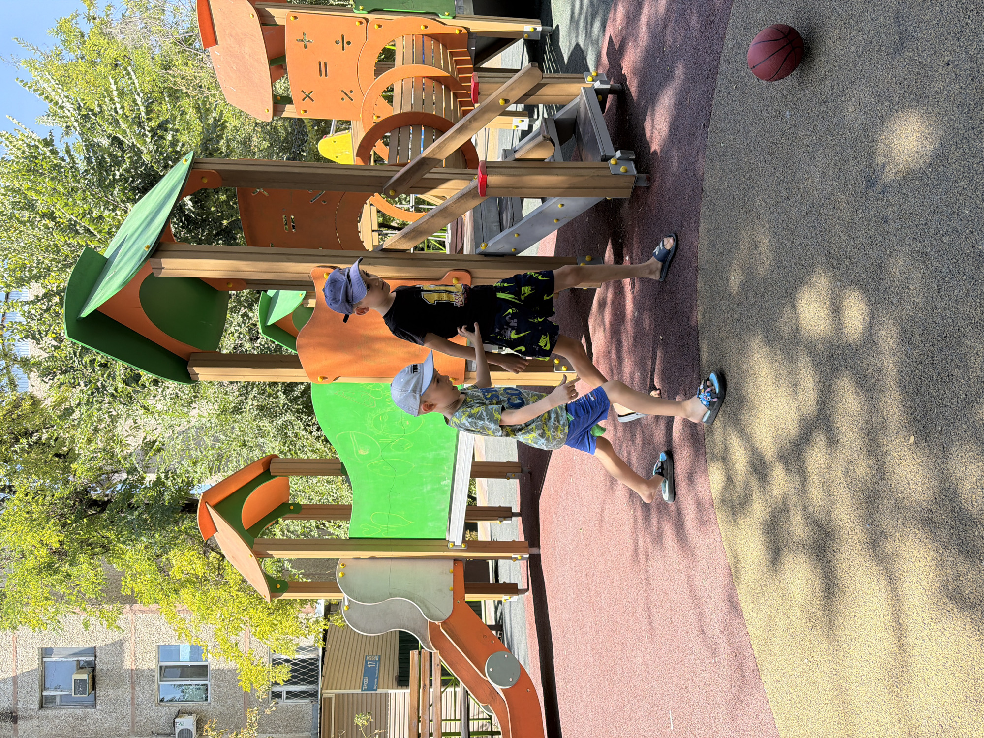 The image depicts a brightly lit outdoor playground scene under a clear blue sky, with lush green trees providing dappled shade. In the foreground, two young boys, both wearing baseball caps, are engaged in activity. The boy on the left is wearing a patterned t-shirt and blue shorts, while the boy on the right is dressed in a black t-shirt with yellow lettering and black shorts with a neon green pattern. They appear to be walking or playing, with their stances suggesting movement. A red basketball rests on the yellow-and-red patterned ground near the lower right corner of the image. The playground structure, made of wood and brightly colored plastic in orange, green, and beige, features slides, stairs, and various interactive elements. In the background, a multi-story apartment building with windows and air conditioning units is visible, suggesting an urban setting. The overall environment is that of a residential area.