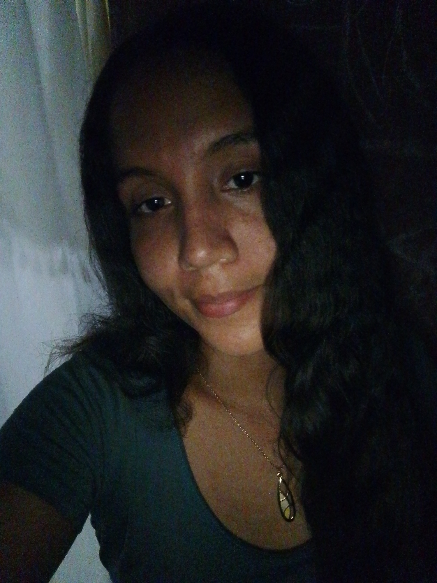 A close-up selfie depicts a dark-haired woman with medium complexion and visible freckles. She is wearing a dark green or blue-green t-shirt and a gold necklace with a teardrop-shaped pendant. Part of a white curtain is visible in the upper left of the frame. The woman's hair is long and wavy, primarily situated on the right side of the image. The lighting is dim, contributing to the shadows in the scene. No specific features indicating the location as Juan Griego, Venezuela, are visible in the photograph. There is no interaction between the woman and other entities in the shot; she is the sole subject, posing for the selfie. The background is largely obscured by darkness.