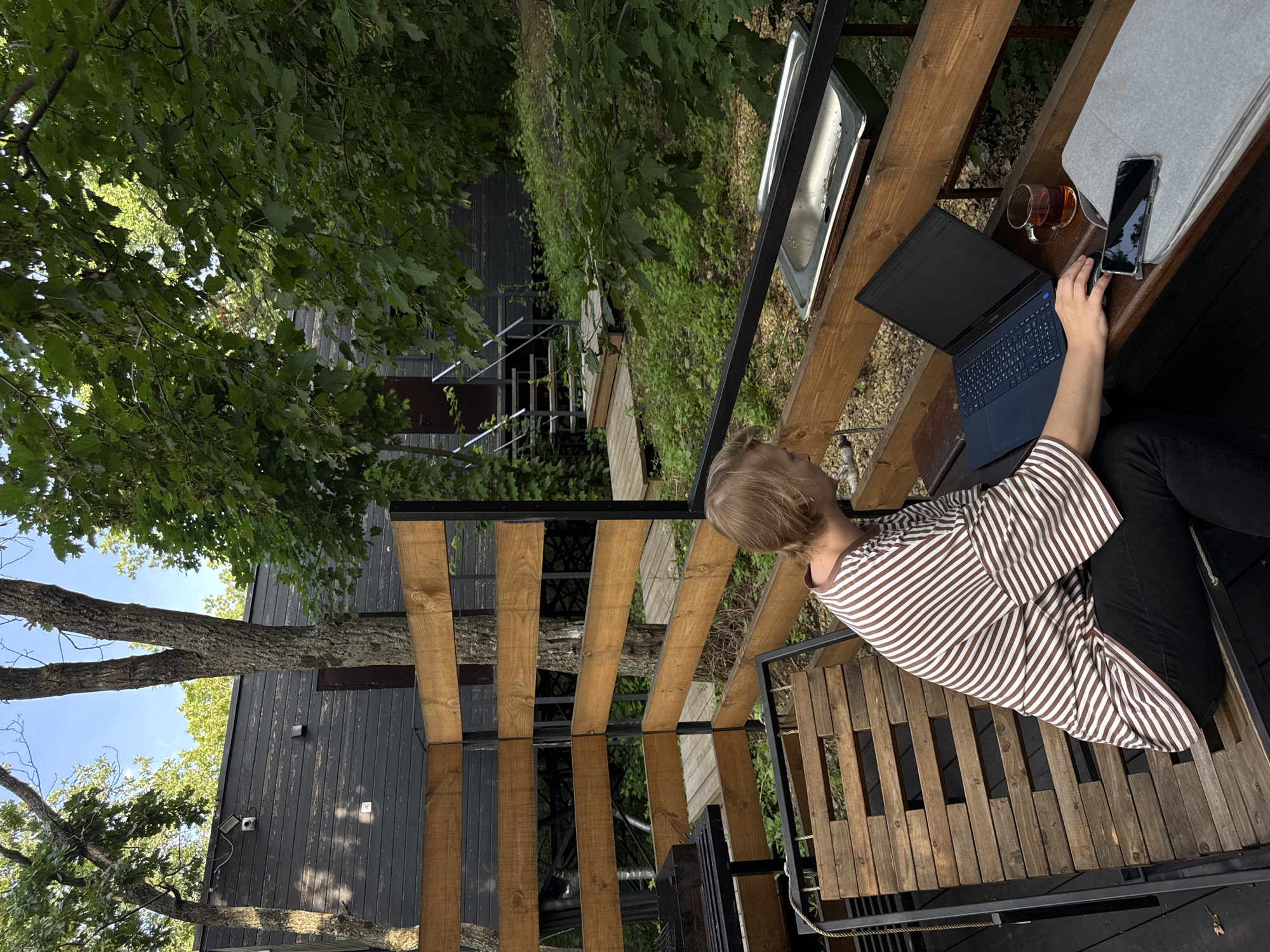 The media file shows a person working on a laptop at an outdoor wooden table. The person, with blonde hair, is wearing a brown and white striped shirt. On the table, there is a laptop, a glass of tea, and a smartphone. The setting appears to be a natural, outdoor environment, possibly a balcony or deck overlooking a wooded area. In the background, there is a dark-colored building and trees. The scene suggests a relaxed, work-from-home or remote work scenario. The lighting indicates it is daytime, and the presence of leaves on the trees suggests it is likely spring or summer. The location context provided is Усть-Курдюм, Russia. There is no visible text in the image.