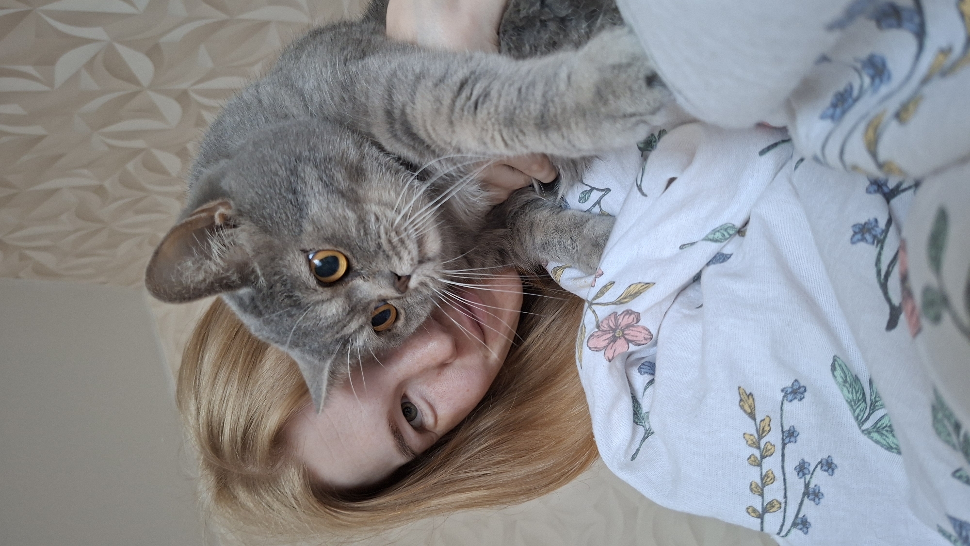 A person with blonde hair, fair skin, and light-colored eyes is holding a grey cat with large orange eyes. They are positioned close to the camera, with the person's face partially obscured by the cat. The person is wearing a white garment with a floral pattern. The background consists of textured wallpaper and appears to be an indoor domestic setting. There is no indication of the specific location within the city of Chaikovsky, Russia.