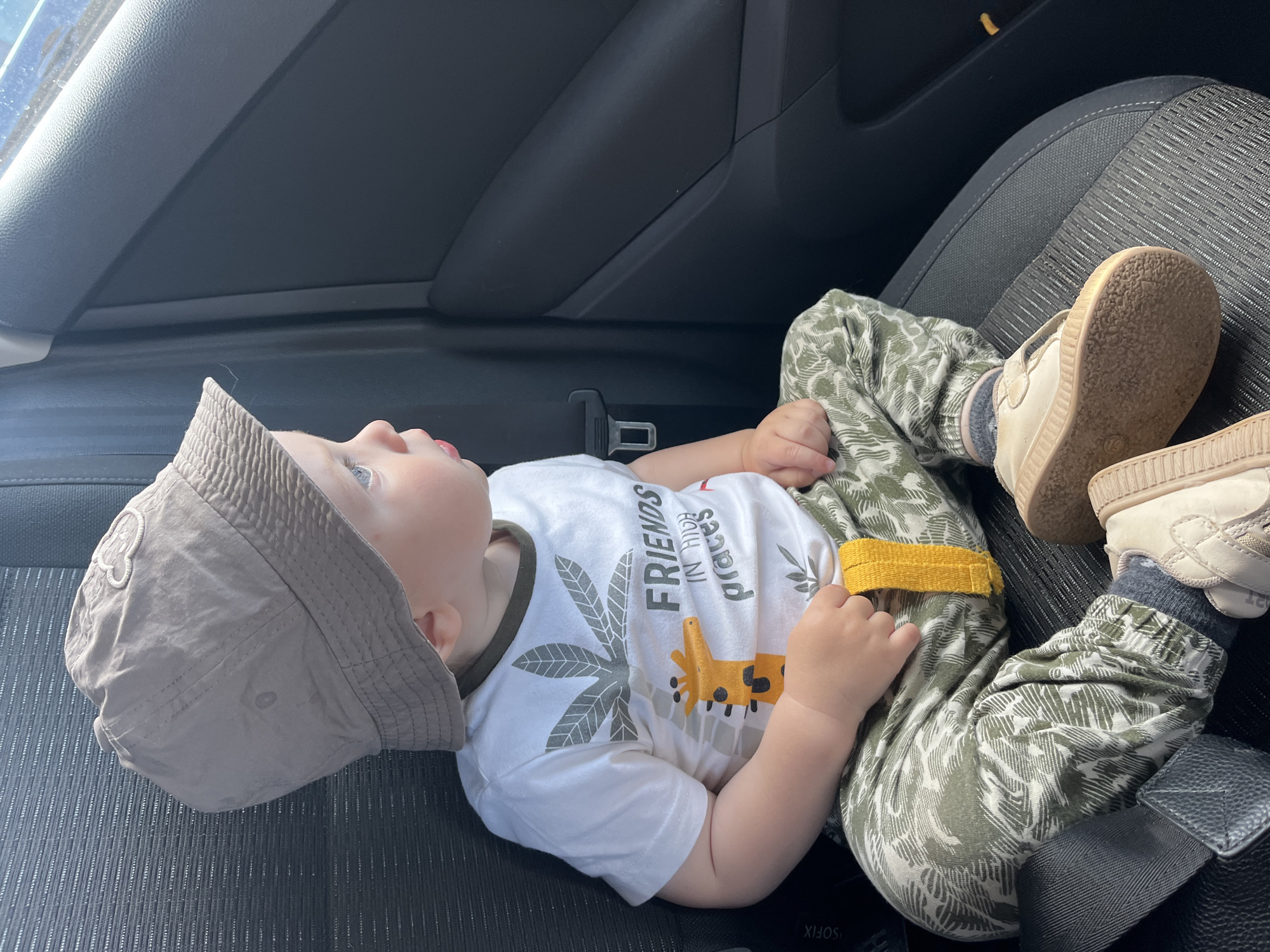 A young child, appearing to be a toddler, is seated in the rear passenger seat of a modern vehicle during what appears to be daylight. The child is wearing a beige bucket hat with a light-colored, possibly embroidered or appliquéd, emblem resembling a cartoon animal head on the front-left side. Their blue eyes are wide and focused intently towards the car's window on the right side of the frame. The child is dressed in a white short-sleeved t-shirt with a dark green trimmed collar. The t-shirt features a graphic design of green-grey palm leaves, an orange giraffe's head, and the text "FRIENDS IN HIGH places" (with "places" partially curved). They are wearing olive green and white patterned jogger-style pants with elasticized ankles, and a yellow fabric loop is visible on the left leg. Grey socks are worn with light beige, low-top shoes featuring light brown soles and Velcro closures; the right shoe is off the child's foot and rests in front of the left foot. The child is sitting on a dark grey textured fabric car seat, and a black seatbelt buckle is visible near their left hip. An ISOFIX label is present on the left side of the seat. To the right, the car's interior door panel, composed of dark grey textured plastic, frames a bright exterior view through the window, showing what appears to be a blue sky or water. There are no discernible landmarks or contextual elements within the image that would identify the location as the city of Chaikovsky, Russia, or any specific geographic area.