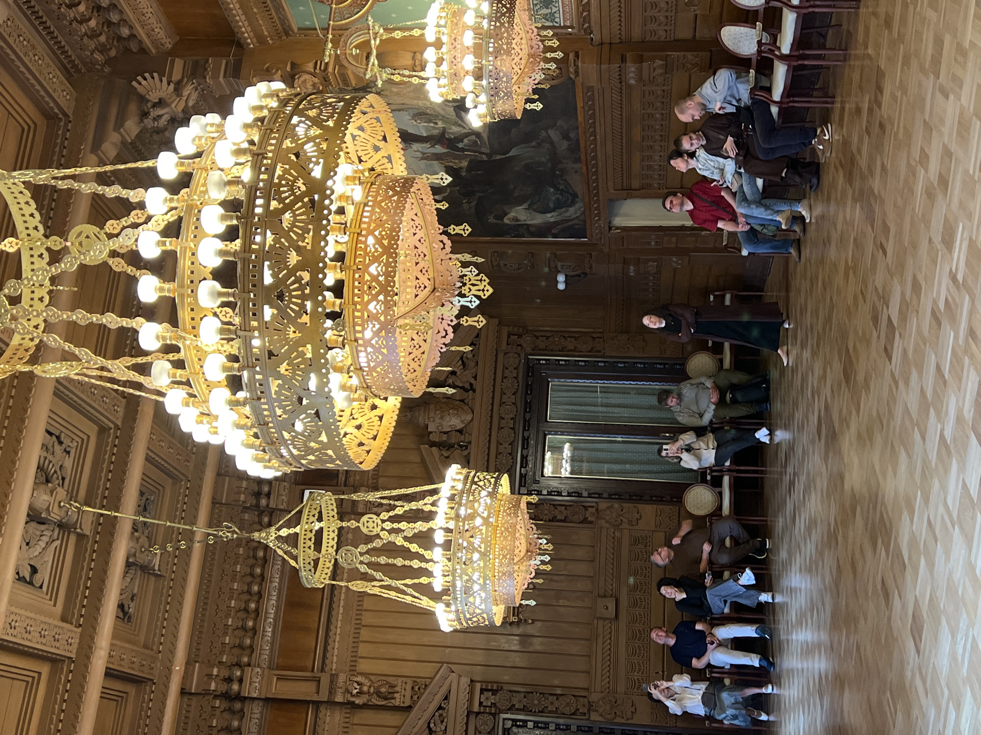 In a grand hall in St. Petersburg, Russia, a group of people are seated on chairs facing a woman standing in a long dark dress and dark boots. The hall features ornate wooden paneling and intricately designed golden chandeliers hanging from the ceiling. The floor is made of polished parquet. The people are of various ages and ethnicities, dressed in casual attire. Most of them are looking up, presumably at the chandeliers or a point of interest above. The woman standing appears to be addressing the seated individuals. One person in the seated group is holding a phone, seemingly taking a picture or recording. The overall atmosphere suggests a cultural event or a guided tour within a historic building.