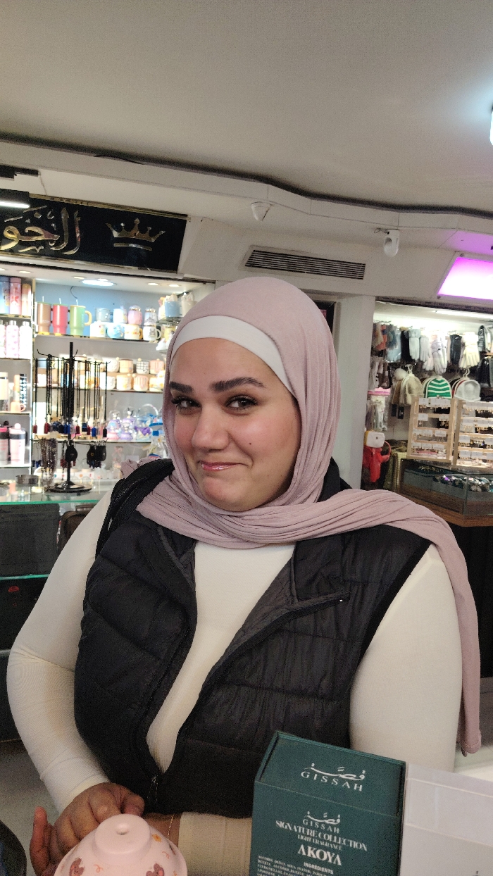 The image displays an adult female in an indoor retail establishment, located in Barza Municipality, Syria. The woman is centered, depicted from the chest up, wearing a light pink hijab over a white undercap, a cream-colored long-sleeved shirt, and a dark grey or black puffer vest. Her face shows a slight smile with glossy lips. Her right hand rests upon a small, light pink decorative object with subtle patterns. To the lower right, a dark green box labeled "GISSAN SIGNATURE COLLECTION AKOYA" is partially visible. The background reveals a retail environment with shelves displaying diverse merchandise, including colorful mugs, various decorative items, black prayer beads, and apparel such as hats and accessories. Overhead lighting includes bright panels, one with a pink hue, and two white dome security cameras are visible on the ceiling. A black sign with gold Arabic calligraphy, "الجوهرة" (Al-Jawhara), and a crown emblem is affixed to the wall behind her.