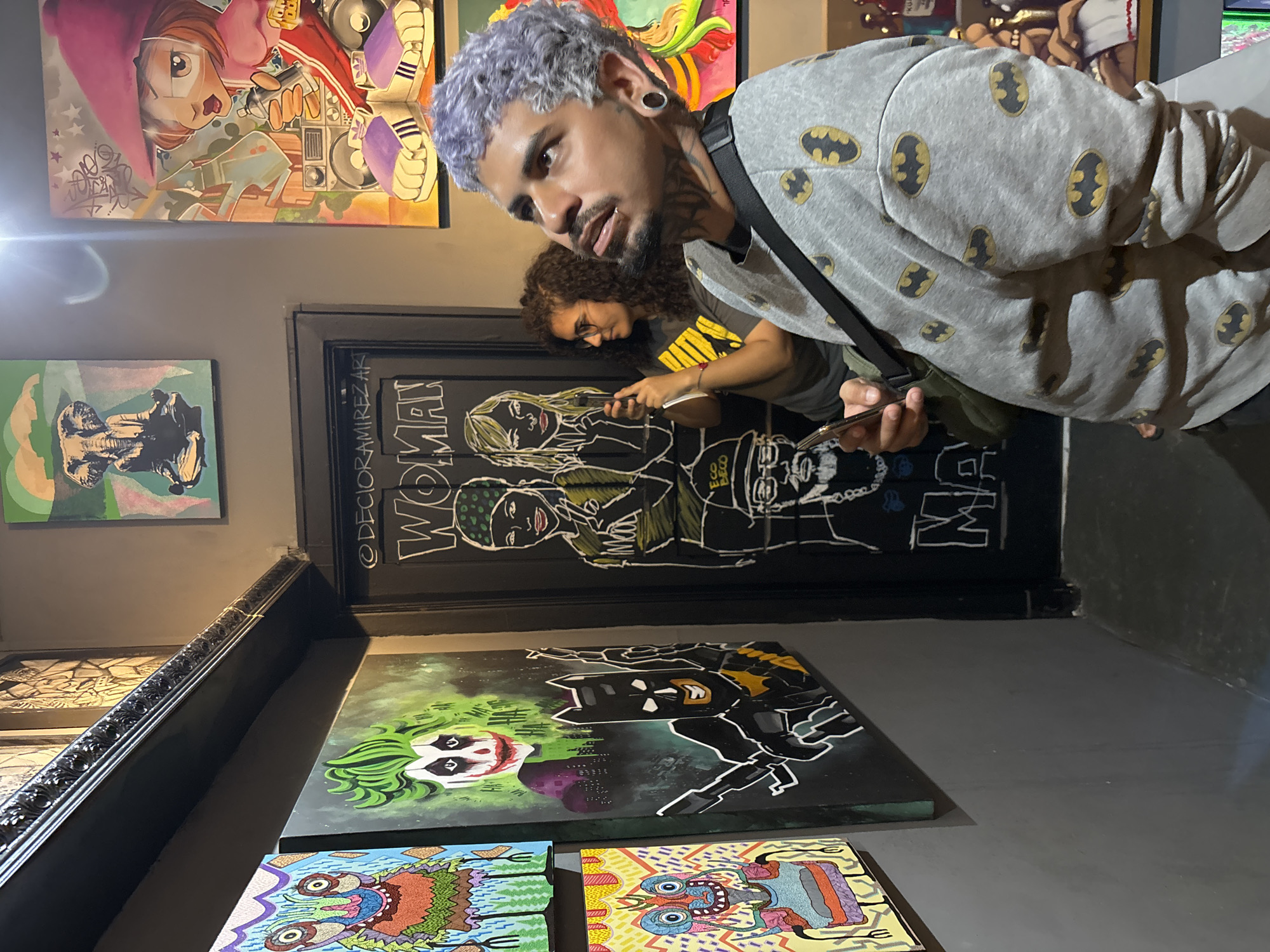 A brightly lit art gallery in São Paulo, Brazil, features artwork displayed on the walls and shelves. A man with dyed grey hair and a Batman-themed shirt stands in the foreground, looking down at his phone. Another person is visible in the background, also looking at a phone. The artwork includes colorful, cartoonish images and some pieces depicting Batman and the Joker. The environment is clean and modern, with a grey floor. The gallery displays both framed and unframed artwork.