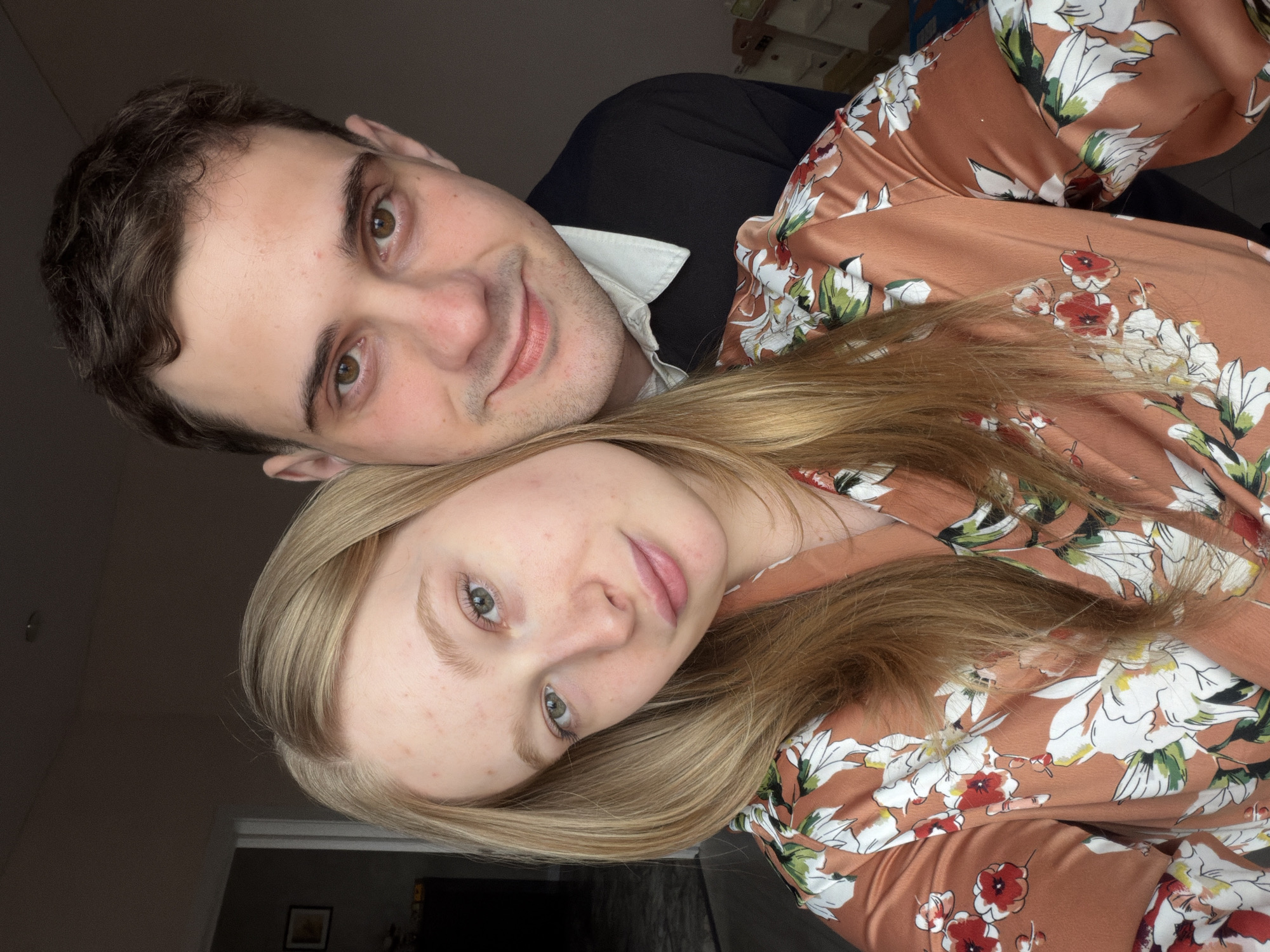 A man and a woman are posing for a selfie indoors. The man has short, dark brown hair, fair skin, and is wearing a black suit jacket over a white collared shirt. He is positioned behind the woman, looking down at the camera with a slight smile. The woman has long, light blonde hair and fair skin with some freckles. She is wearing a floral-patterned robe or top with a brown background and white, red, and green flowers. Her face is tilted towards the camera.
The background is dimly lit, with a glimpse of a doorway and a framed picture on the wall in the distance. There are also some objects visible in the background that appear to be part of the interior decor.