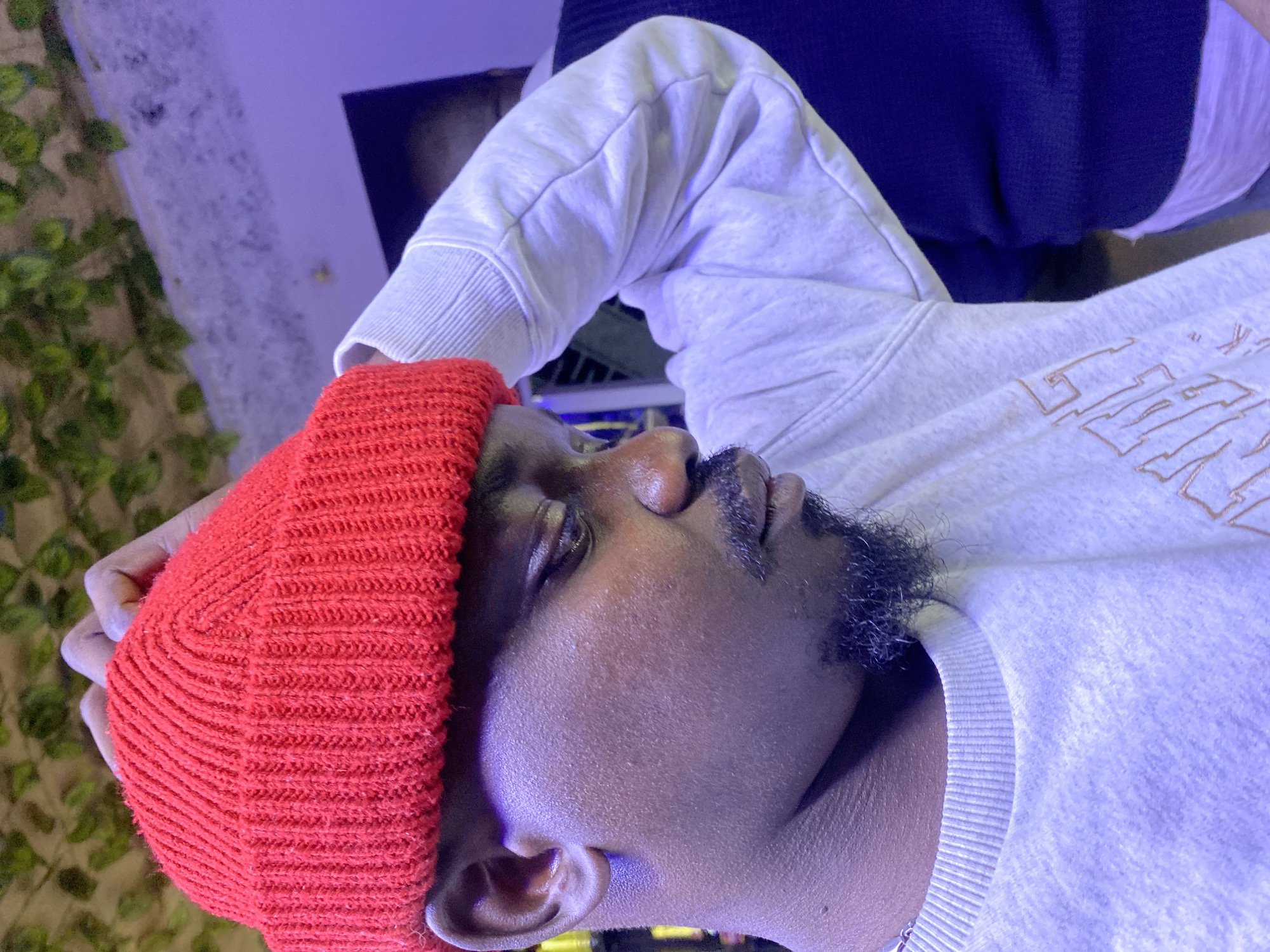 A dark-skinned male is depicted in a close-up, three-quarter profile, adjusting a bright red knit beanie with his right hand. He wears a light grey long-sleeved crewneck sweatshirt with a faint graphic design, revealing a white undershirt underneath. His head is tilted downwards, and his gaze is directed to his right. He has a moustache and a short, dark goatee. The scene is illuminated by strong purple ambient lighting, casting a prominent hue on the subject's skin and clothing, as well as the immediate background. To the upper left, a section of green foliage is visible against a neutral-toned wall. In the right background, a second individual wearing a dark blue ribbed sweater over a white undershirt is partially visible. The setting is an indoor establishment located in Bar Jirgi Summa, Nigeria.