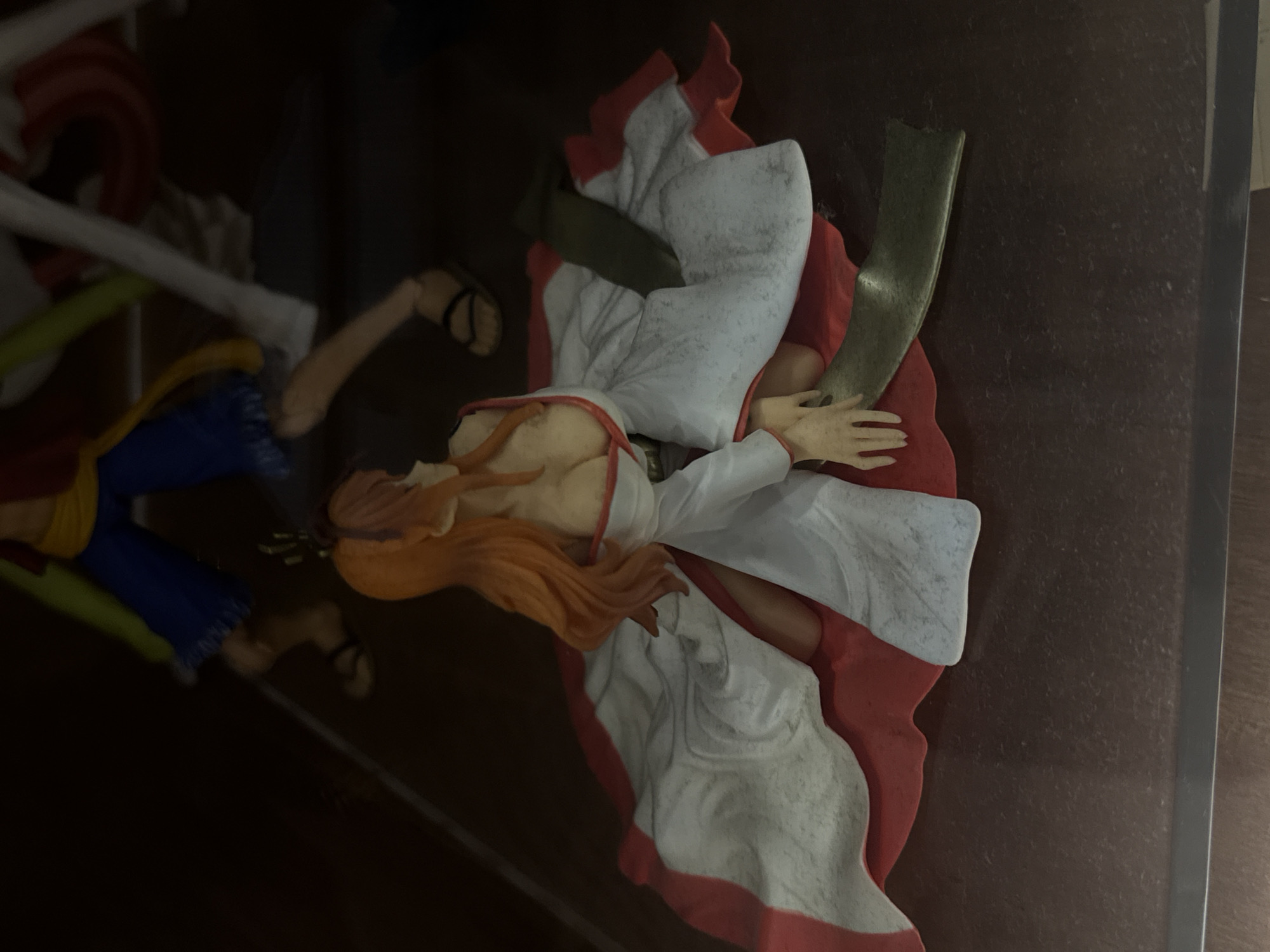 The image shows a close-up, low-angle shot of a Nami figurine from the anime series One Piece. The figurine is detailed and appears to be made of plastic. Nami is depicted kneeling on a dark, textured surface, wearing a white and red kimono. Her orange hair is visible, and she is looking away from the camera. The background is dark and out of focus, with hints of other figures or objects. The lighting is dim, creating shadows that add to the atmosphere. The figurine's pose and attire suggest a narrative or thematic representation.