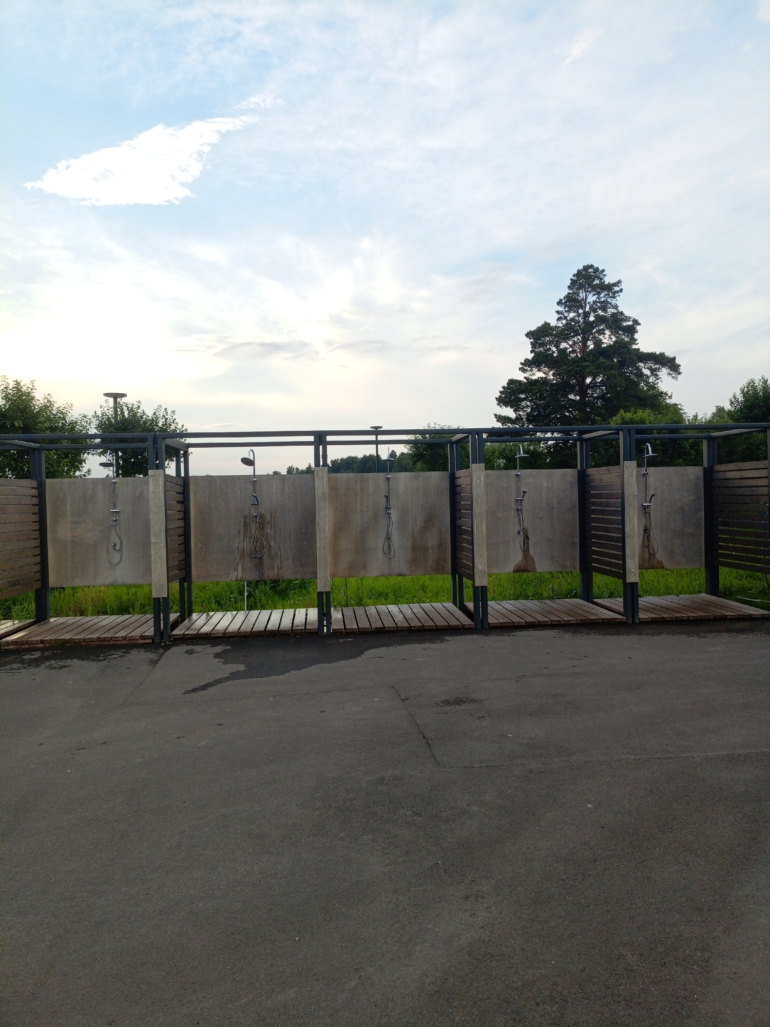 The image shows an outdoor shower facility with four individual shower stalls, each featuring a shower head and faucet. The stalls are constructed with a metal frame, wooden slat walls on the sides, and concrete back panels. Wooden planks form the flooring within each stall. The area is paved with asphalt in the foreground, and a patch of green grass and vegetation is visible behind the shower stalls. A large pine tree stands prominently in the background, with a cloudy sky above. The overall scene appears to be a public amenity, possibly located at a campsite or recreational area in Zheleznogorsk, Russia. There are no people visible in the image, and no actions or interactions are taking place. The lighting suggests it is daytime, with overcast conditions.