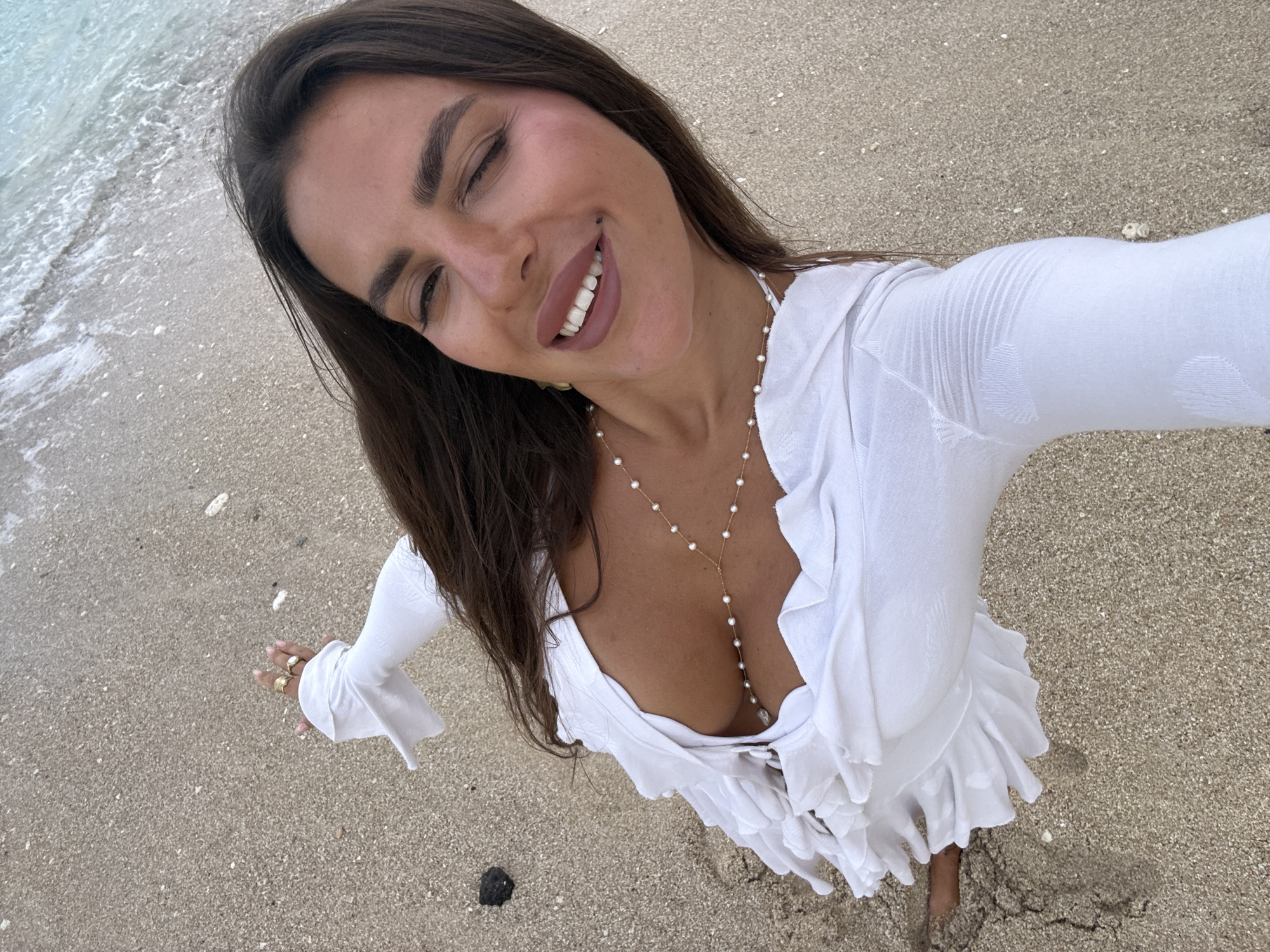 A woman with long, dark hair is taking a selfie on a sandy beach. She is wearing a white, ruffled dress with a plunging neckline and a pearl body chain. Her left hand, adorned with rings, is raised and touching her hair, while her right hand holds the camera. The ocean waves are visible in the background. The scene is set in Kuta Selatan, Indonesia.
