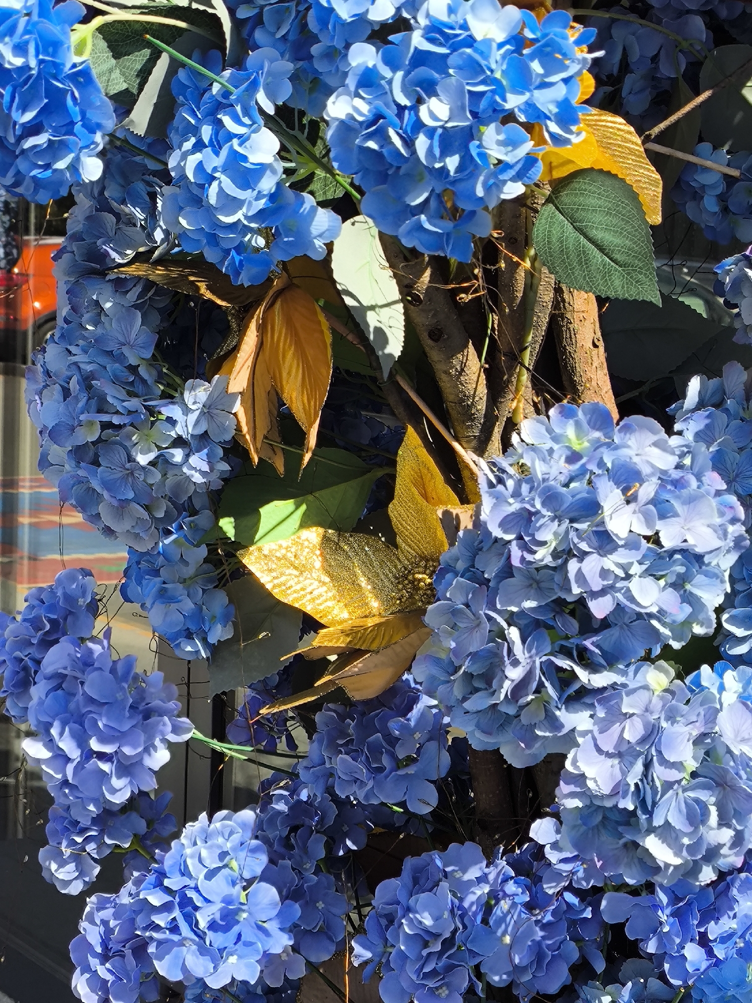 A close-up, eye-level, outdoor shot features a cluster of artificial blue hydrangeas arranged around a textured, brown, tree-like trunk. The flowers, some with lighter blue and white accents, fill most of the frame, with a few gold and green artificial leaves interspersed. The background shows a blurry reflection of a building with orange and blue stripes, suggesting an urban environment. No people or specific actions are visible. The scene appears to be a decorative arrangement, possibly in a city like Yekaterinburg, Russia, given the provided context.