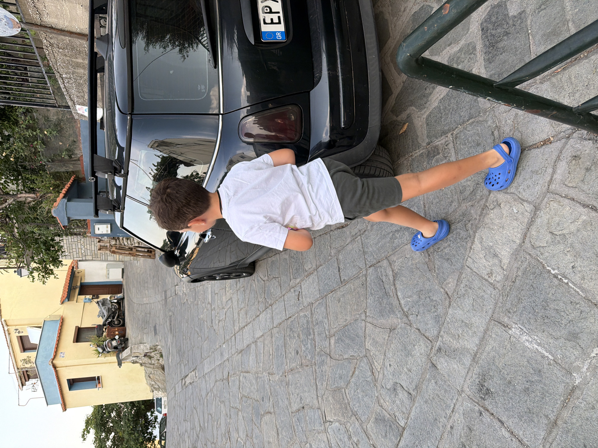 A boy wearing a white t-shirt and gray shorts walks on a cobblestone street in Kavala, Greece. He is wearing blue Crocs. To his right is a black Mini Cooper with a roof rack and a Greek license plate. In the background, there is a yellow building with blue trim and two parked motorcycles. The scene appears to be a residential street.