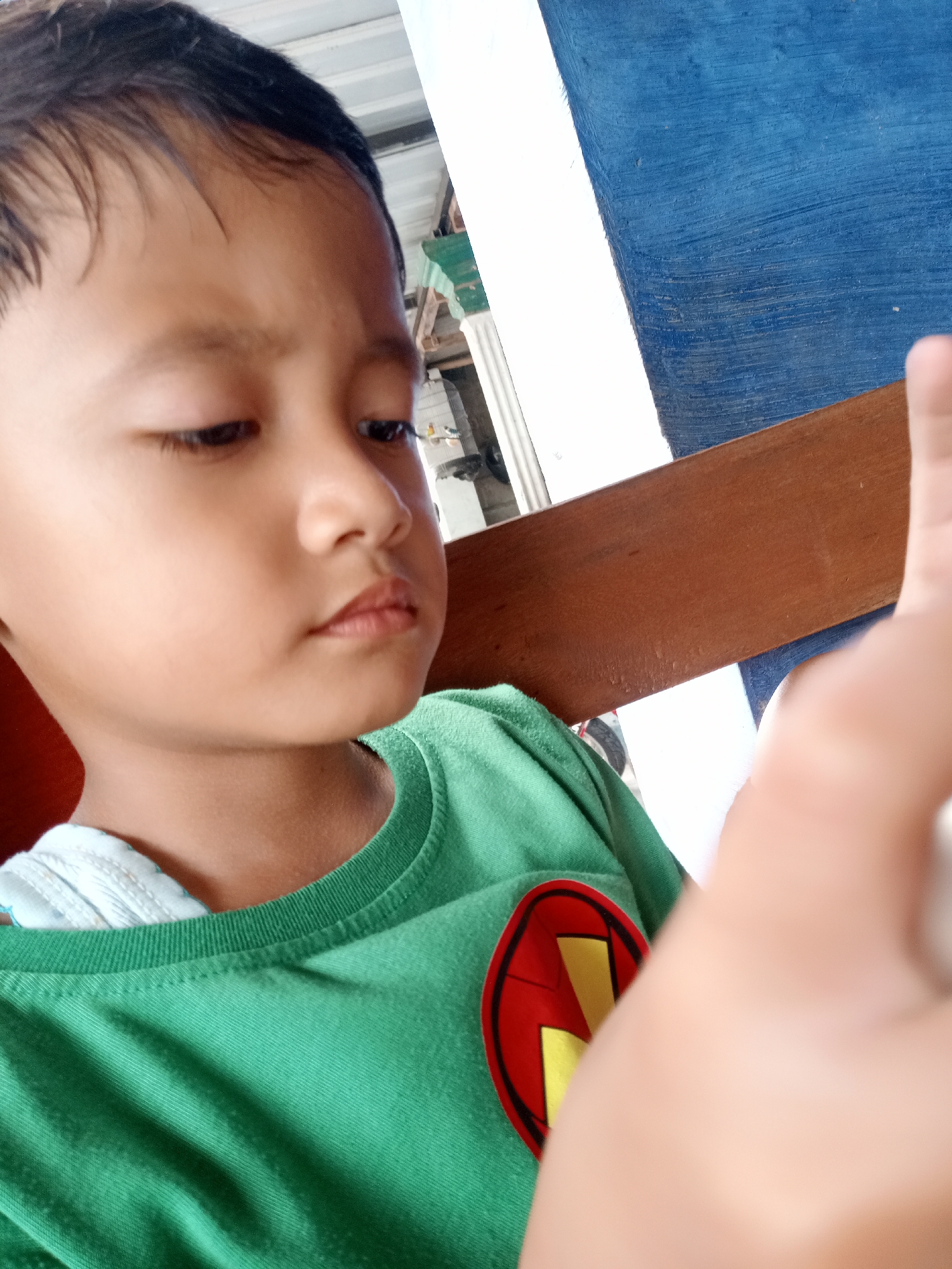 A close-up, eye-level shot features a young child, likely a boy, focused downwards and slightly to the right. The child has short, dark hair and a concentrated, neutral expression, suggesting engagement with an unseen object or activity. He is wearing a bright green short-sleeved t-shirt with a red circular emblem on the left side of the chest, featuring a yellow, stylized design resembling a 'W' or 'M'. A light blue undergarment is visible at the neck of his t-shirt.
The background includes various surfaces: a textured blue wall or partition is visible on the right, positioned above a horizontal brown wooden beam or railing. A white vertical surface runs adjacent to the blue wall. Further to the left and in the upper background, hints of light-colored walls, a ceiling, or structural elements are present. A blurred hand, possibly an adult's, occupies the lower right foreground, partially obscuring the child's t-shirt and the background, potentially holding the camera or an item the child is looking at.
The scene appears to be set in an indoor or semi-enclosed environment, such as a room, porch, or covered area, during daylight hours. The setting is casual, consistent with a domestic space or local establishment in Bungur, Indonesia. The primary activity is the child's focused attention on something out of frame. No visible text is present in the image.