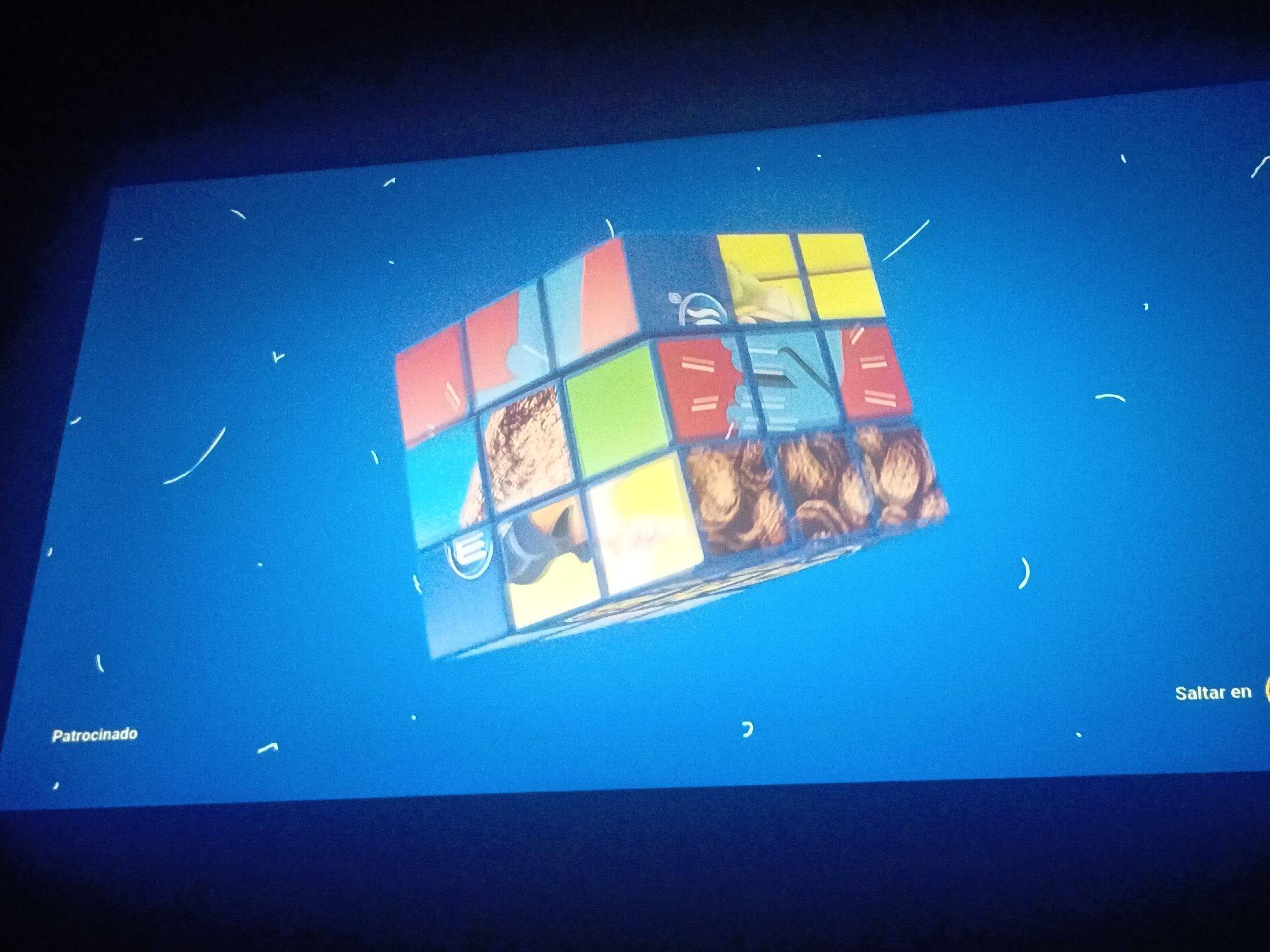 A television screen displays an image of a Rubik's Cube with images of various foods on its faces. The background is a solid blue. The screen displays text in Spanish: "Patrocinado" and "Saltar en". No information from the city of Juan Griego, Venezuela, is contained within this image.