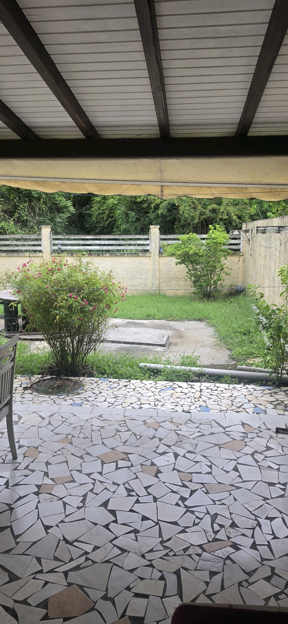 The image depicts a covered patio area with a mosaic tile floor overlooking a backyard. The patio is supported by wooden beams and a yellow awning. In the backyard, there is a bush with pink flowers, a table with chairs, a patch of grass, a concrete slab, and a fence. Beyond the fence, there is dense greenery. The scene appears to be in Capesterre-de-Marie-Galante, France, based on the provided metadata, suggesting a residential or outdoor dining area.