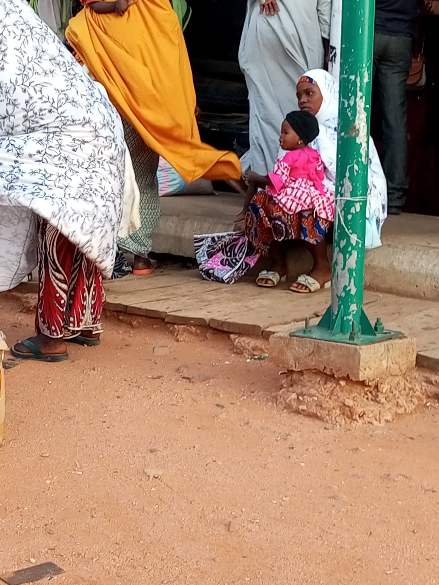 In Gombe, Nigeria, a woman in a white hijab with subtle floral patterns sits on a concrete platform next to a green metal pole. She holds a young child on her lap, who wears a bright pink top and a patterned skirt. The child has a black head covering. Behind them, other individuals are visible, one wearing a large orange garment draped over their shoulder. The foreground features a reddish-brown dirt ground with some scattered debris. The scene appears to be outdoors, possibly near a building, given the presence of the concrete platform and the pole.