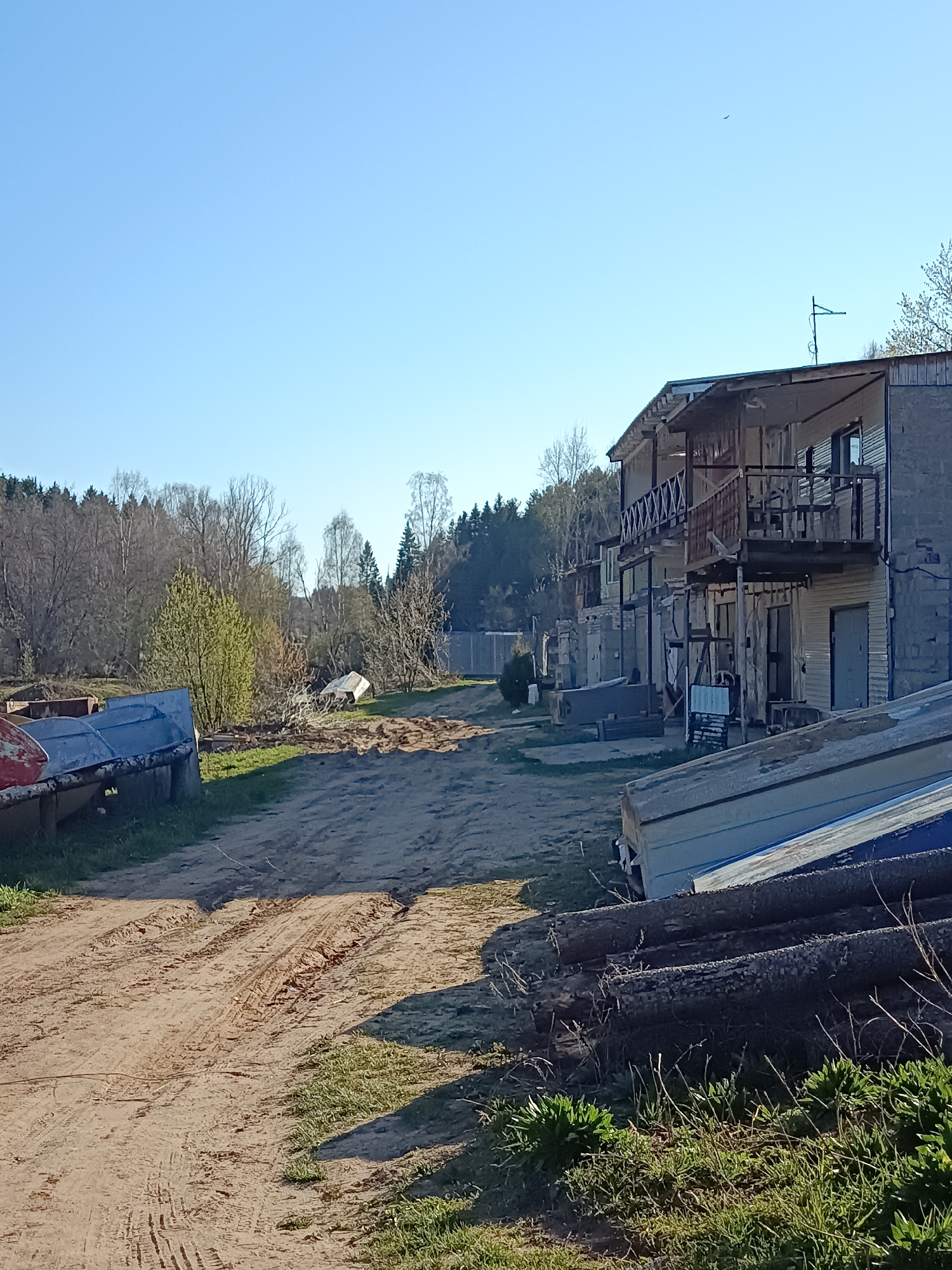 The image depicts a dirt road in what appears to be a rural or semi-rural area in Chaikovsky, Russia.
The road is uneven with visible tire tracks. On either side of the road are various boats, some overturned, indicating possible proximity to a body of water. There is also a pile of logs on the right side of the road. A two-story building with a balcony runs along the right side of the frame. The building appears weathered.
In the background, there is a wooded area with trees that are mostly bare, suggesting early spring. The sky is clear and blue.