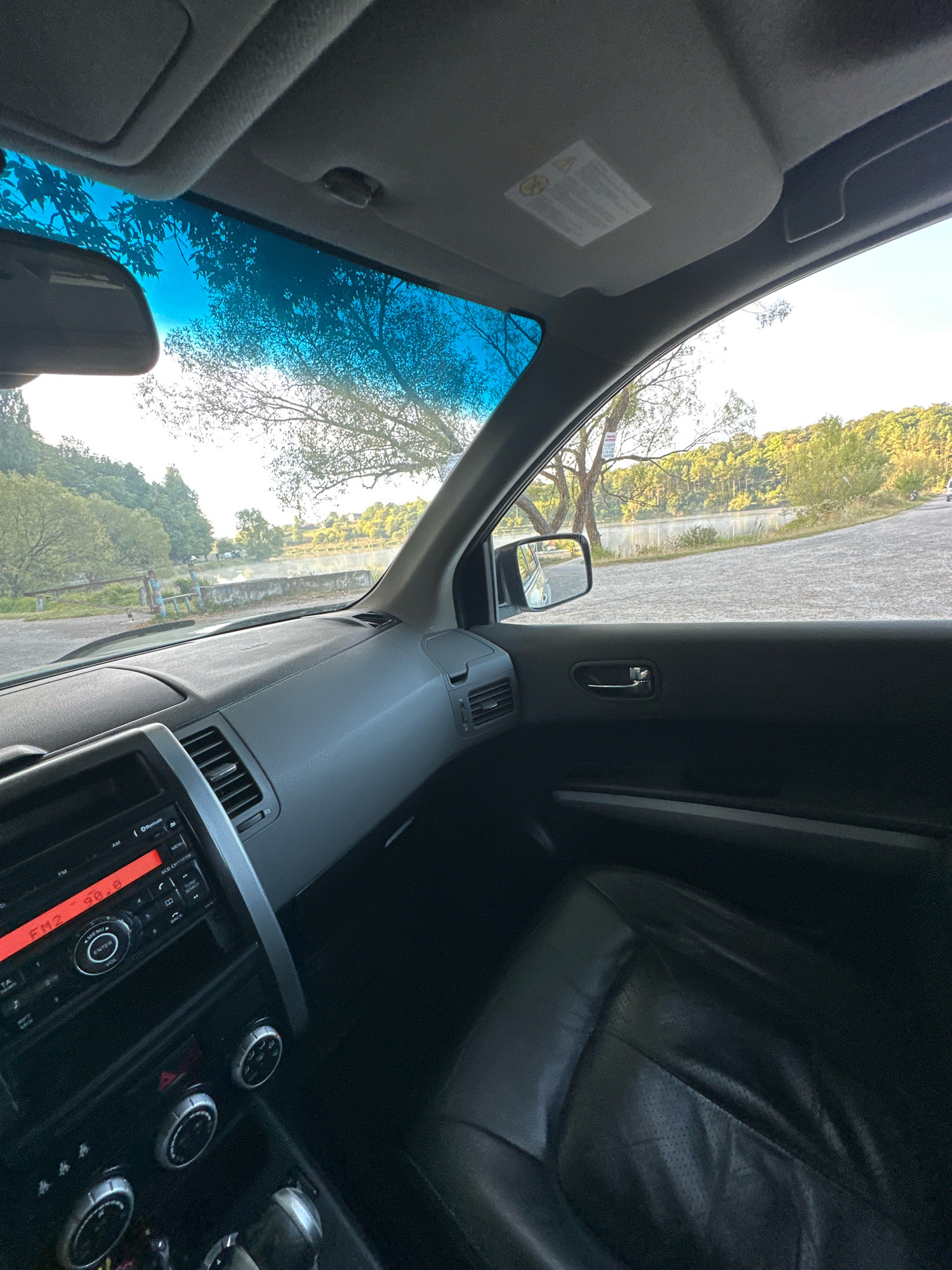 The image is taken from inside a car, looking out through the windshield and driver's side window at a body of water and trees. The interior of the car features a dashboard with a radio and climate controls, a black leather seat, and a rearview mirror. The exterior view shows a calm body of water, possibly a lake or river, with trees lining the far bank. The sky is bright, suggesting daytime. The scene is set in Makiv, Ukraine, with a road visible leading away from the viewer. There are no people visible in the image.