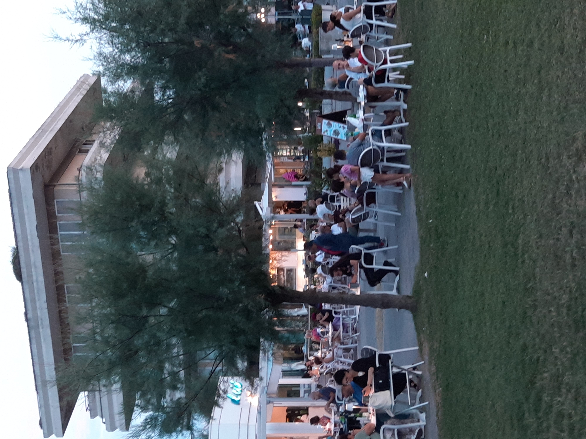 A street view captures a lively outdoor dining area in Pesaro, Italy, with people seated at tables under the shade of trees. The scene features a multi-story building in the background, likely a hotel or apartment complex, with a distinctive architectural style. White tables and chairs are arranged on a paved area, and some tables are adorned with small lamps. Many individuals are engaged in conversation, enjoying meals, or looking at their phones. The foreground is dominated by a grassy area, suggesting a park or public space adjacent to the dining area. The overall atmosphere is one of a relaxed, social gathering in a European coastal town.
